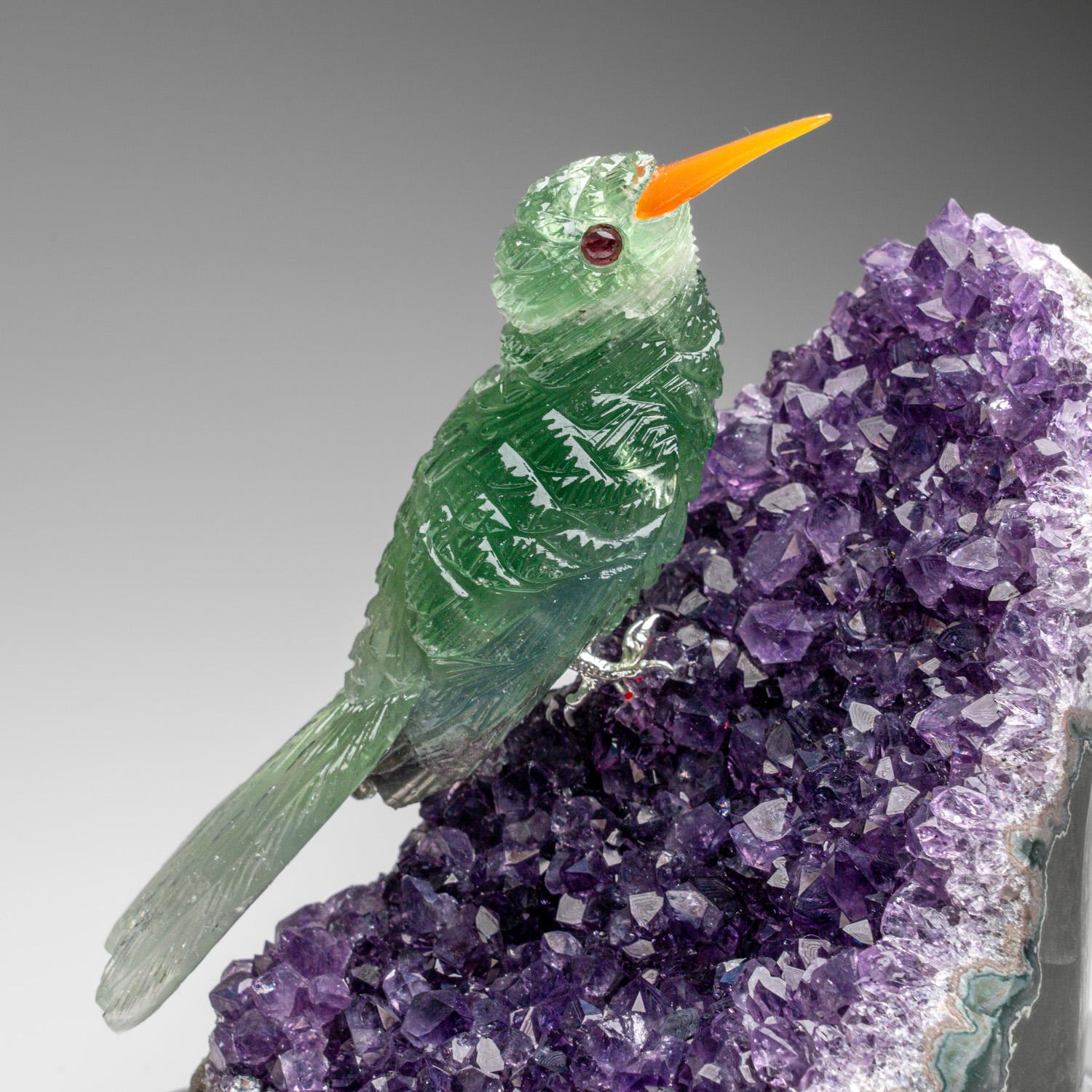 Esta talla de Colibrí es una obra de arte única, tallada en fluorita de calidad de museo, con ojos de granate y pico de cornalina. El colibrí está encaramado sobre una matriz de geoda de amatista y montado en un soporte a medida para exponerlo