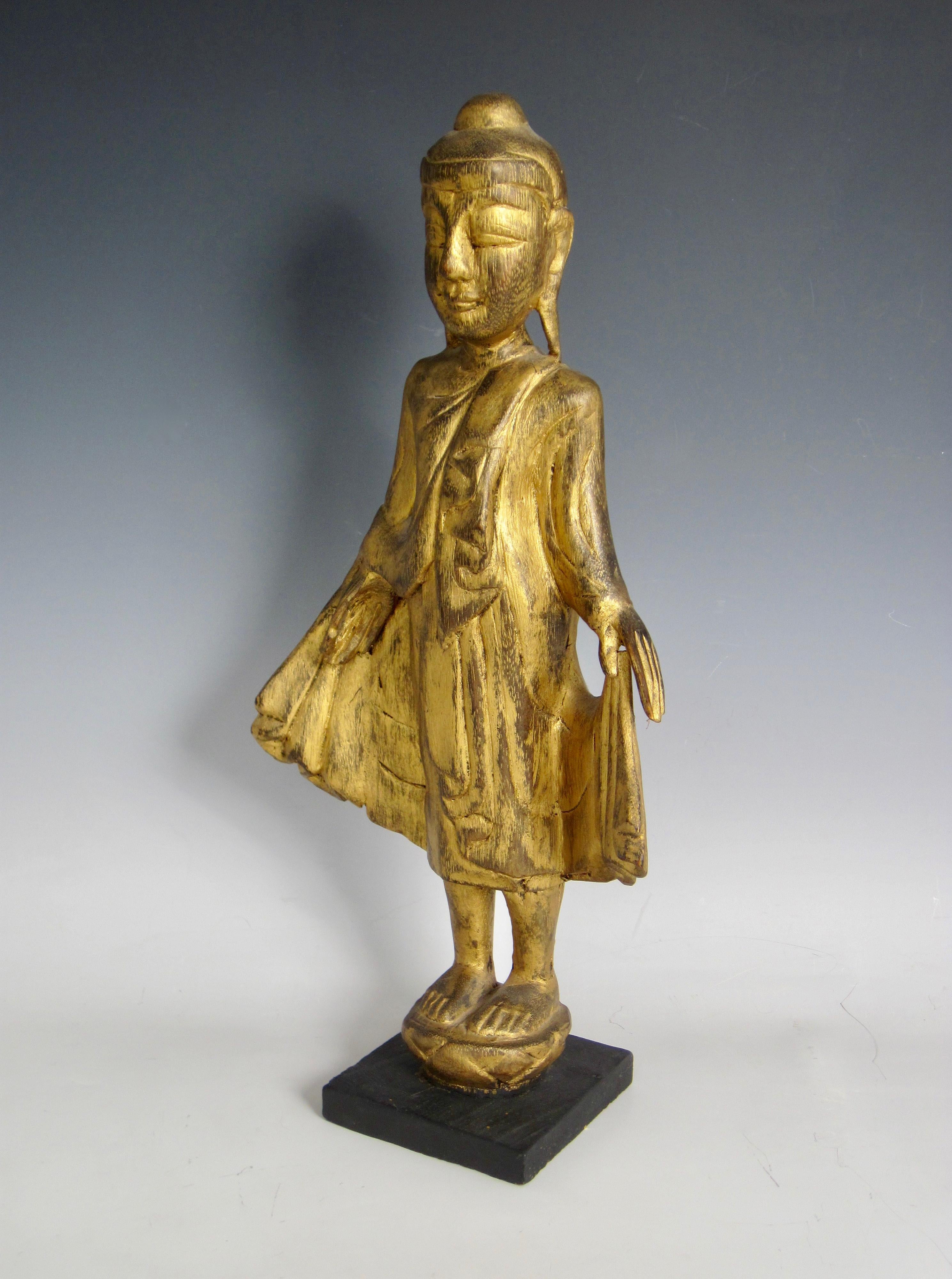 A decorative hand carved Thai giltwood standing Buddha on a lotus flower with black square base. The costume is layered and flowing and the facial expression is peaceful with a grin. Serenity and openness to life is the feeling I get with standing