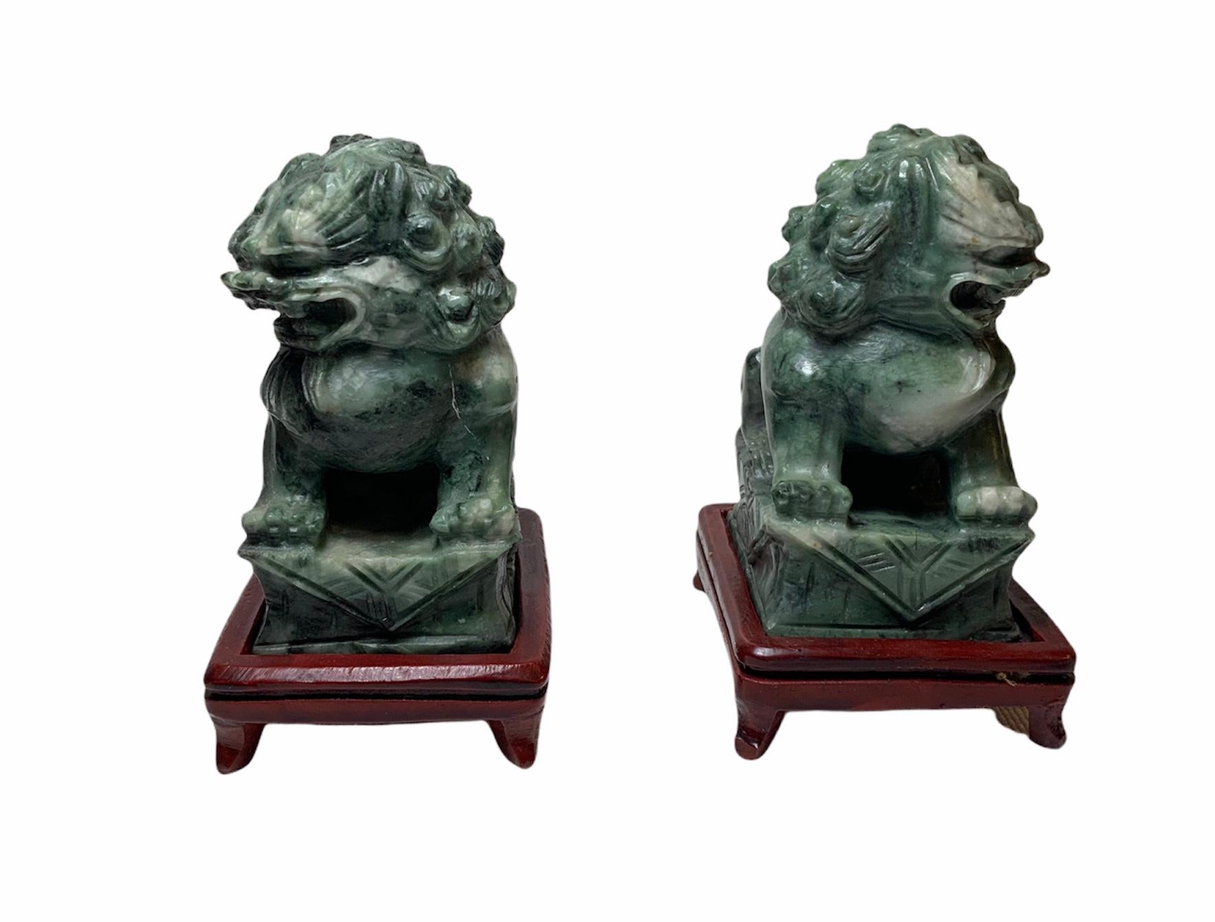 This is a pair of hand carved green stone Chinese Foo-Dog sculptures/figurines. They depict a pair of foo-dogs seated in a rectangular shaped base with a carpet. They are supported by a dark red wood bases with scroll feet.
