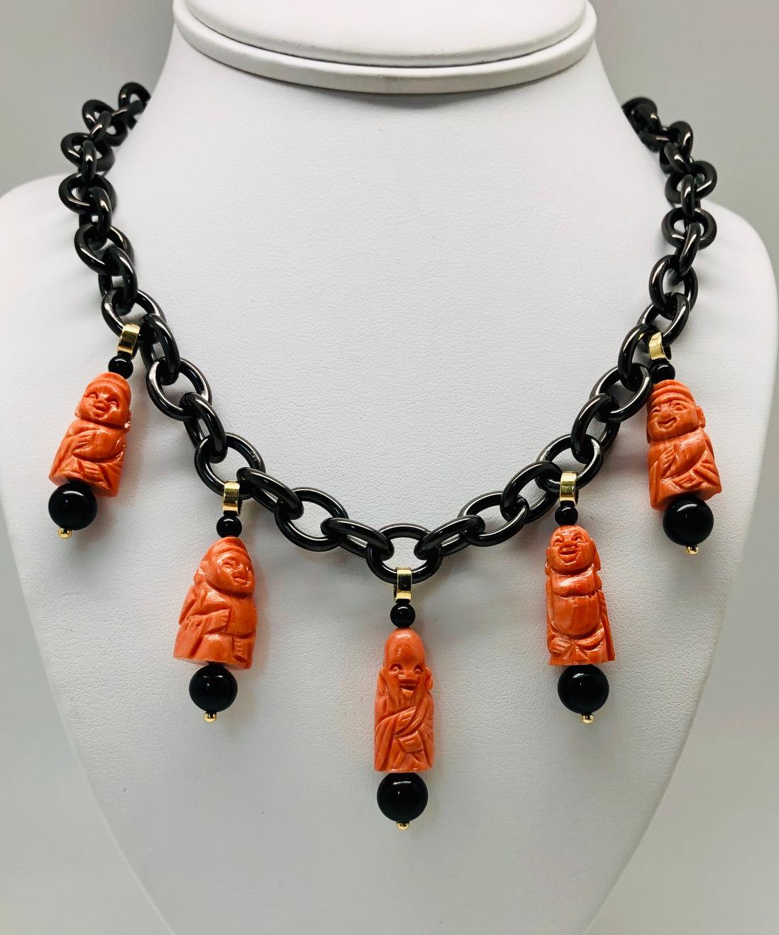 Diese ungewöhnliche Halskette ist aus geschwärztem Stahl gefertigt und besteht aus 5 mediterranen Korallenperlen, die von Hand zu glücklichen Buddhas und Gottheiten geschnitzt wurden. Mit glänzenden schwarzen Onyxperlen und Akzenten aus 18-karätigem