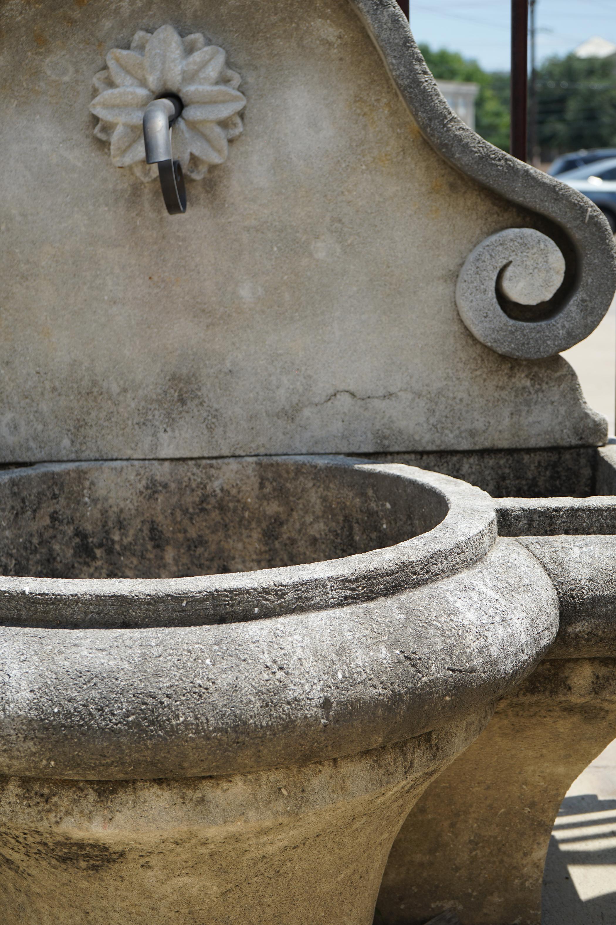 This limestone wall fountain has a circular basin to catch and return the water, but it also has two smaller separate areas carved out to each side for planting seasonal flowers or greenery. The water exit on the back plate has a beautiful lilly