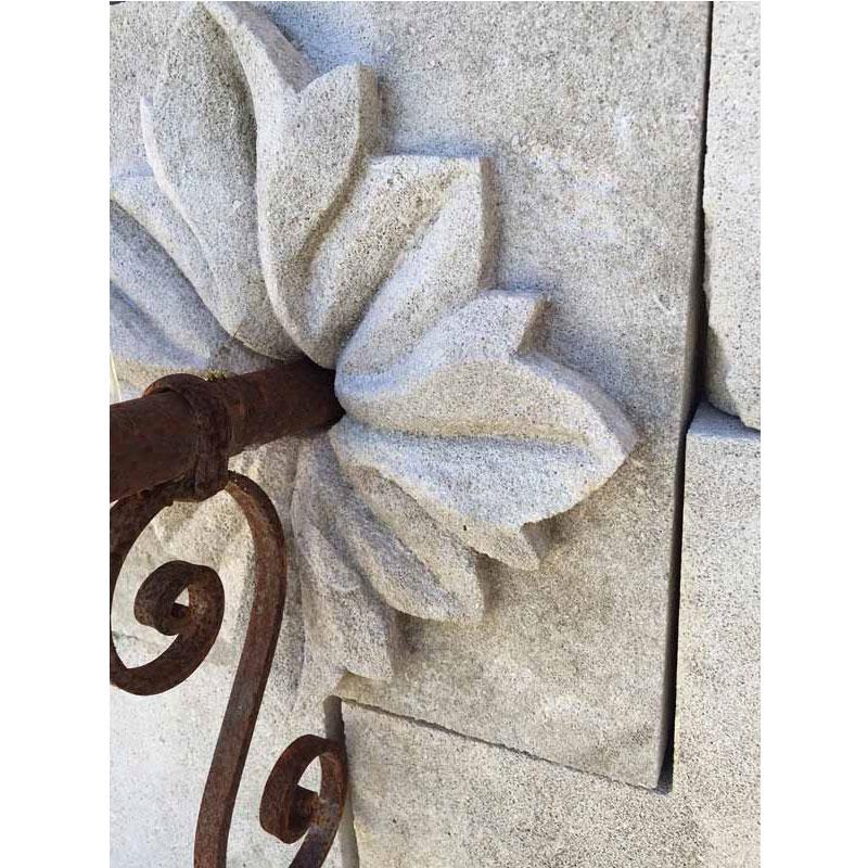 A hand-carved limestone wall fountain from France, featuring an elegant iron spout. Around the spout, a detailed lily carving adds a refined touch, framed by fluted columns on each side. The top center showcases another intricately carved lily,