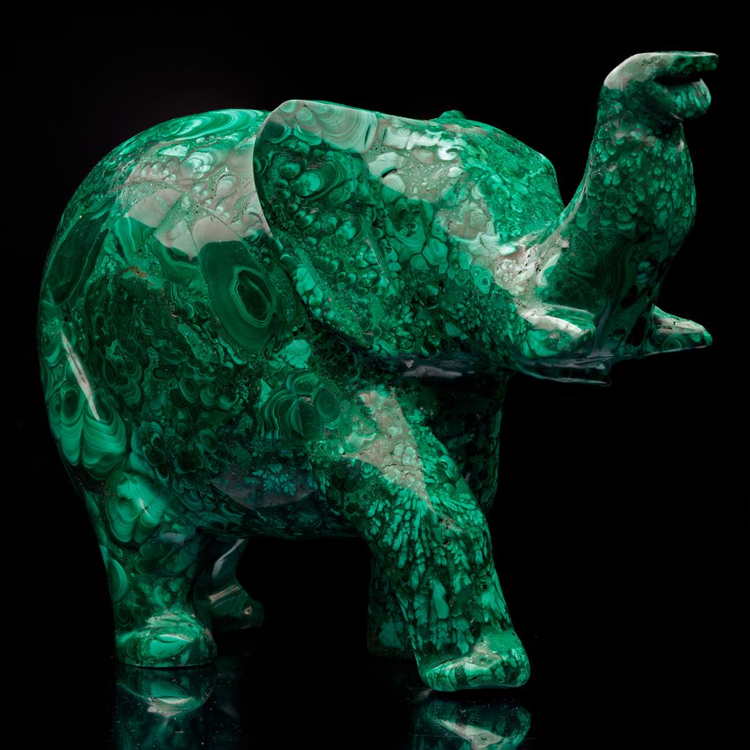This luscious green copper carbonate mineral specimen from the DRC has been expertly hand-carved into a large elephant with upward-facing trunk and hand-polished to a brilliant luster to enhance its gorgeous colors and banding. Malachite was