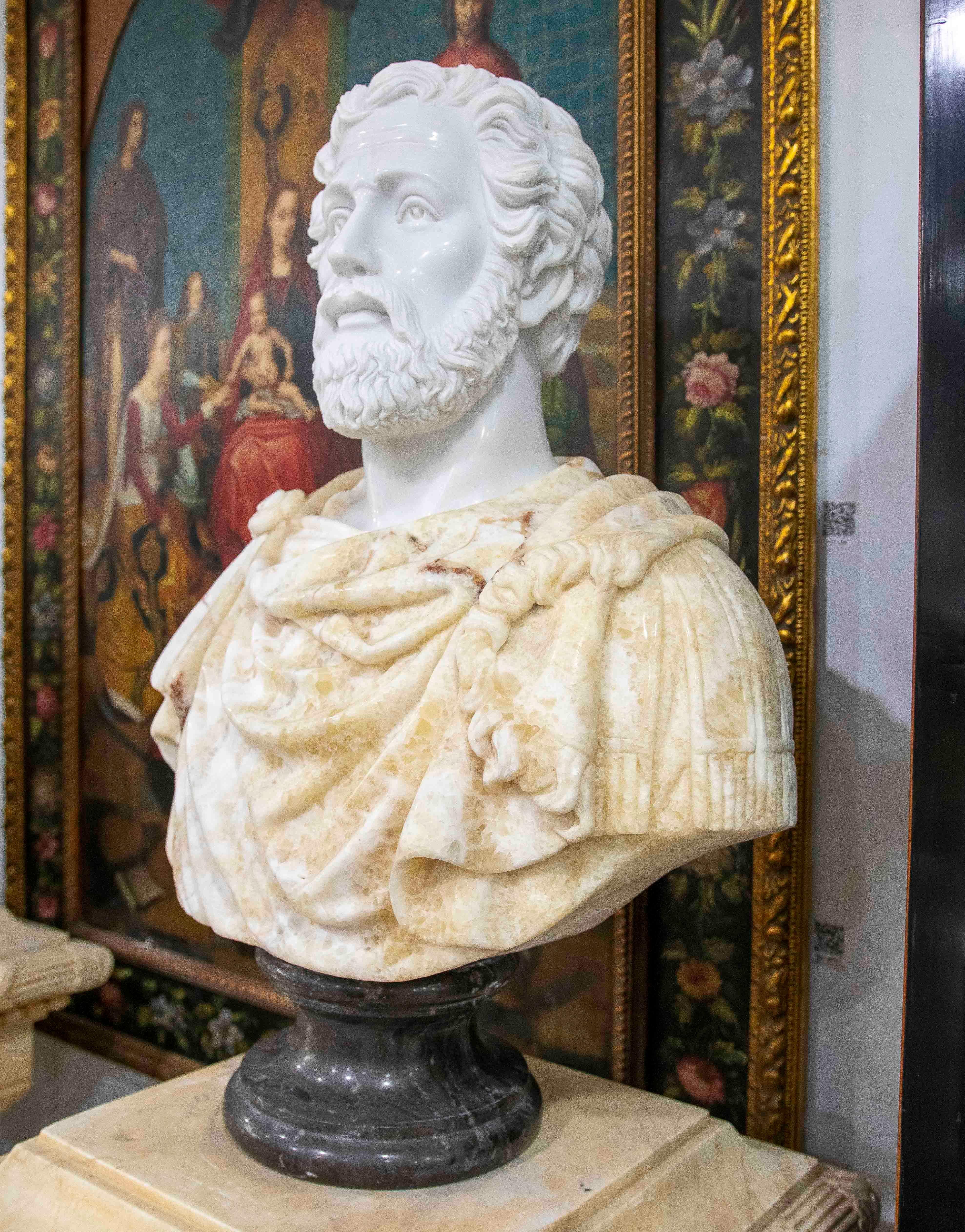 Impresionante busto clásico de mármol, tallado a mano con un detalle excepcional. La pieza representa una figura masculina barbuda con expresión serena y majestuosa, vestida con una túnica romana ricamente elaborada en mármol veteado de tonos