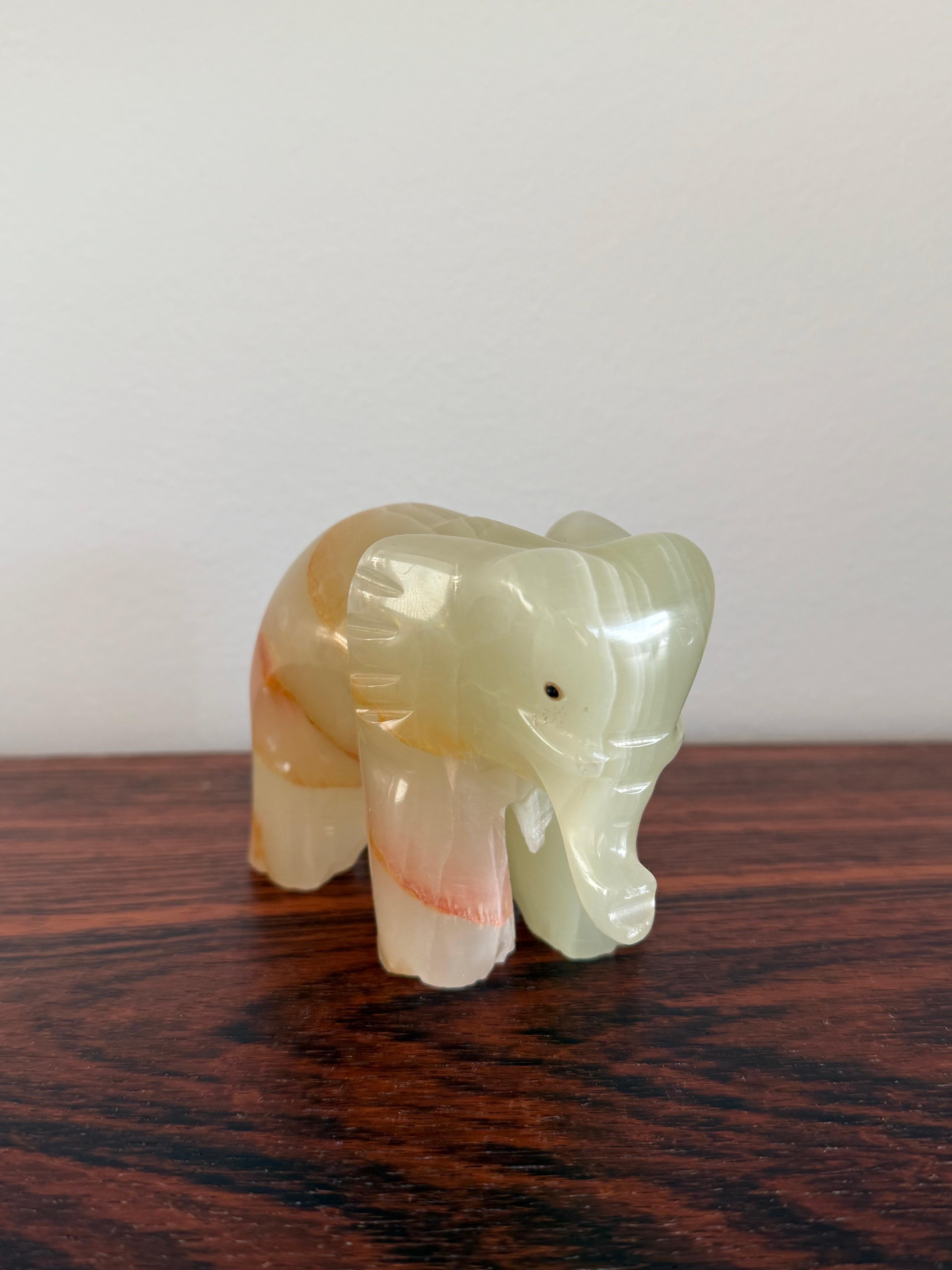 Un'elegante scultura di elefante intagliata a mano in onice naturale, che mette in mostra i luminosi strati di tonalità crema, miele e rosa della pietra. La lavorazione artigianale rivela la traslucenza e la profondità intrinseca del materiale,