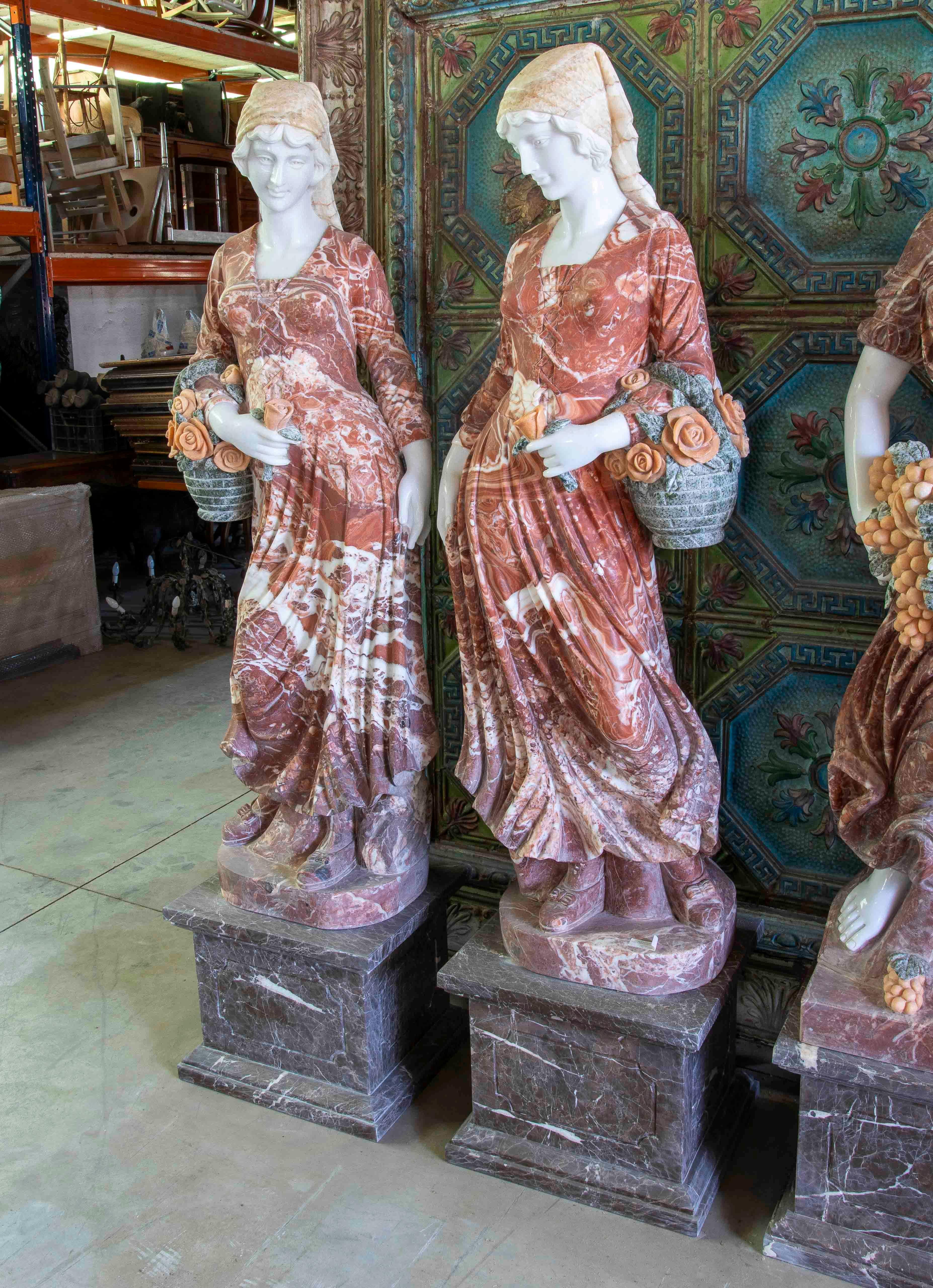 Magnifique paire de sculptures en marbre sculptées à la main représentant deux figures féminines dans une pose sereine, chacune portant un panier de fleurs. Les œuvres se distinguent par la combinaison de différentes teintes de marbre : blanc pour
