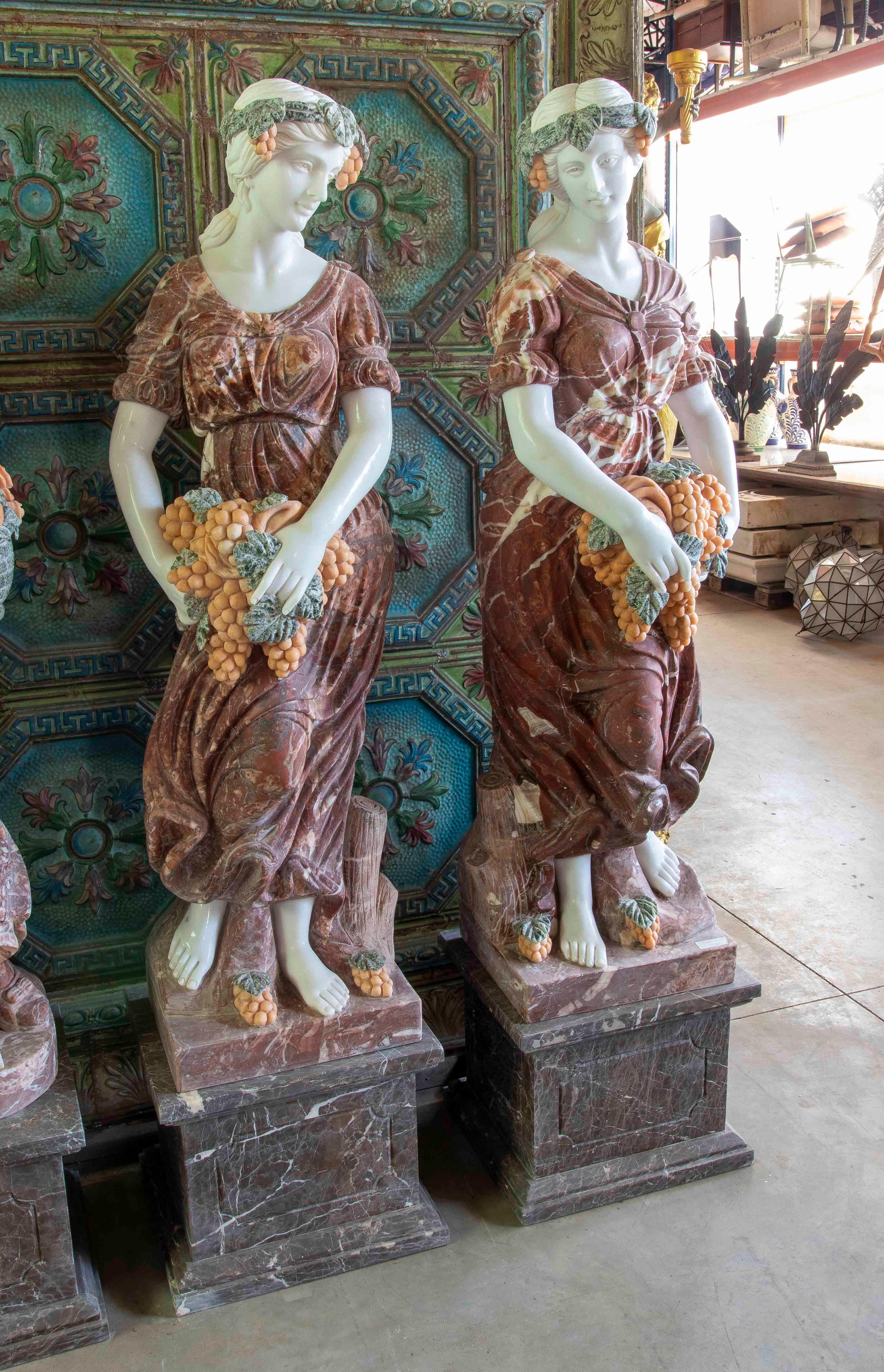 Un ensemble élégant de sculptures en marbre sculptées à la main représentant deux figures féminines classiques portant des grappes de raisin et des feuilles de vigne. Sculptée avec une grande précision, chaque figure combine différents types de