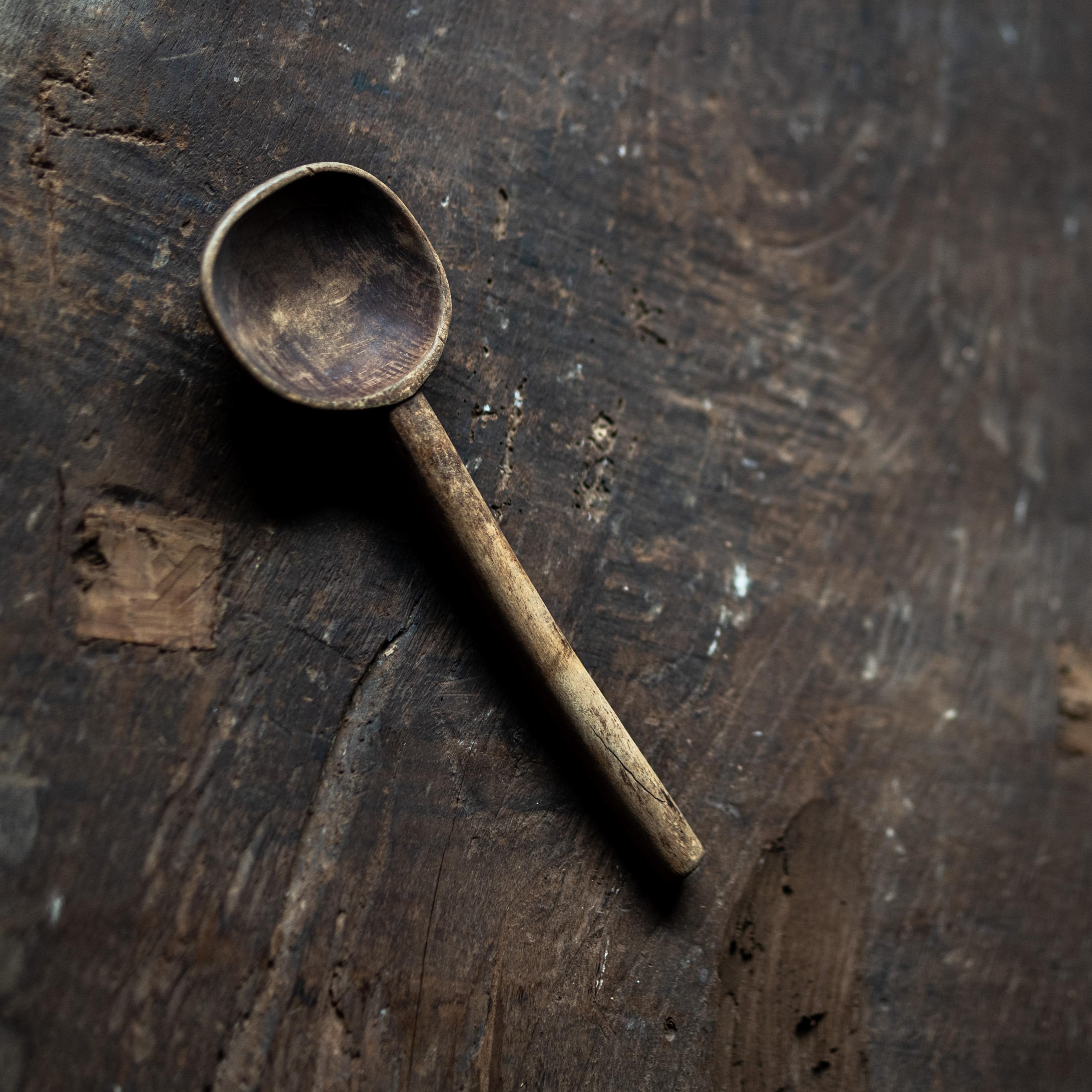 A beautifully weathered hand-carved wooden spoon with a long handle and slightly squared bowl. The object bears the marks of practical use, yet its form and patina give it a sculptural presence. Likely used in a farmhouse or rural kitchen in the