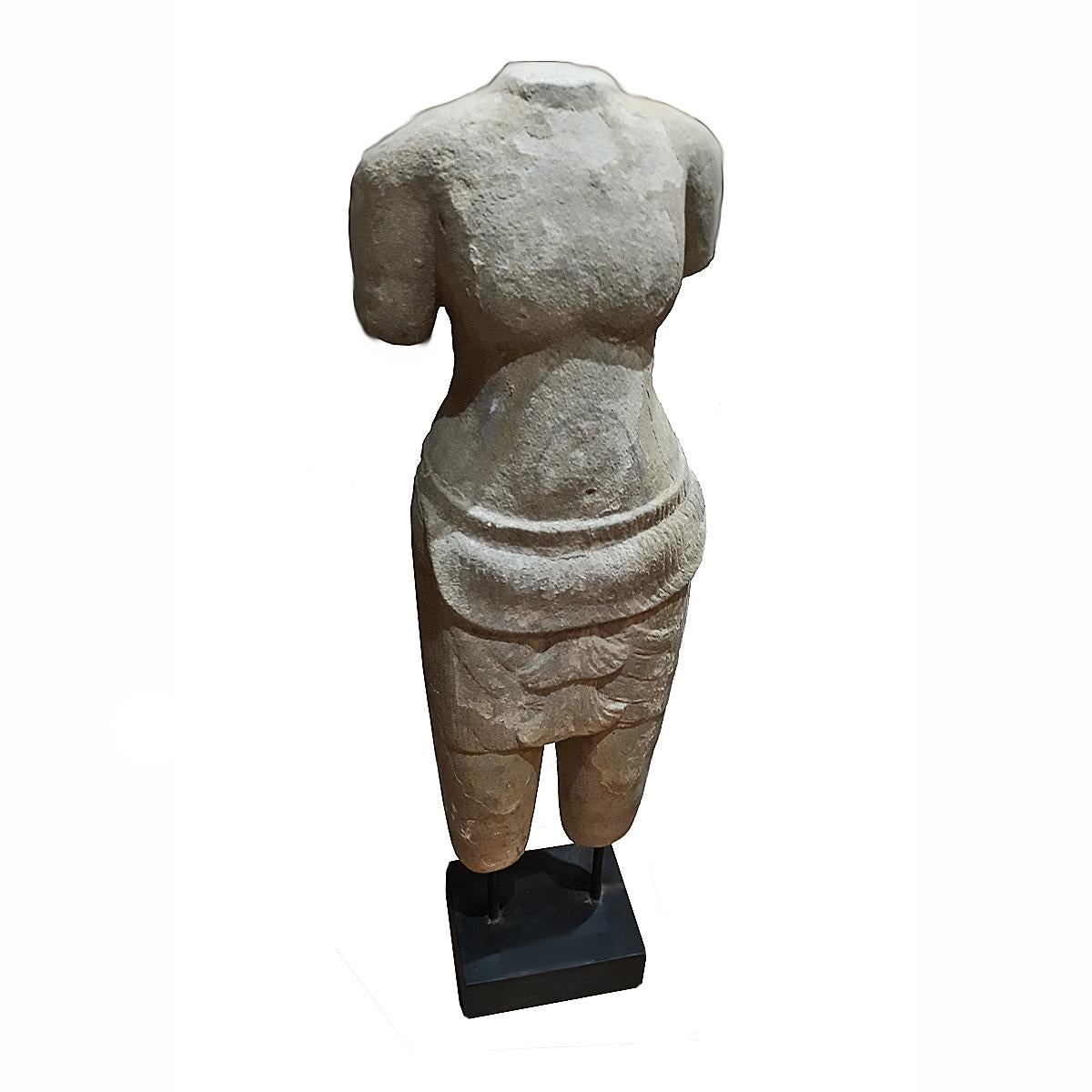 A table-top sculpture of a female torso in traditional attire, hand carved from a single piece of sandstone. From Thailand. Mounted on a wood / metal stand. 29 inches high.