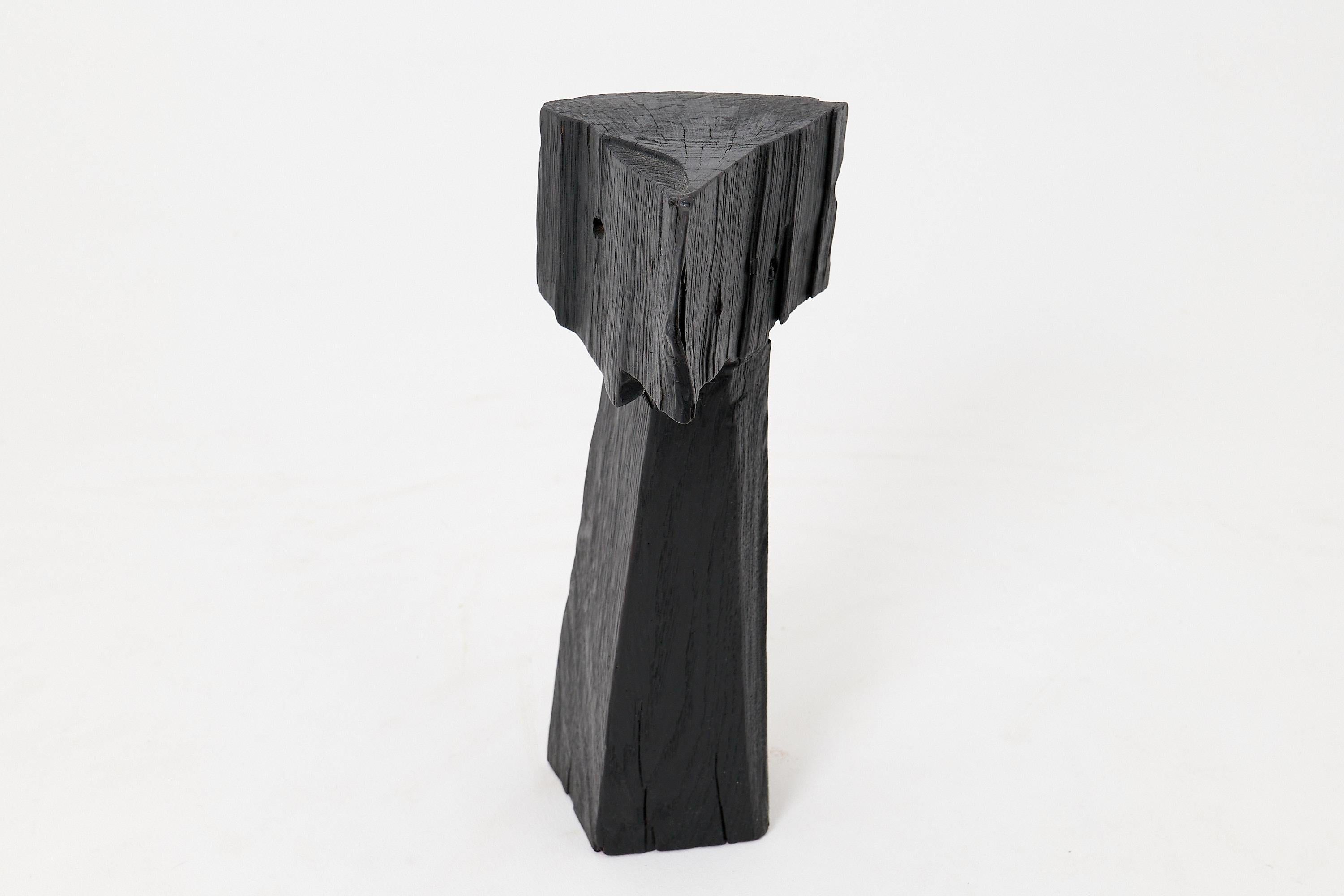 Cette sculpture minimaliste est une pièce unique, sculptée à la main dans un seul bloc de bois dur massif. Sa forme géométrique stricte évoque un totem moderne ou une figure abstraite, définie par un lourd coin angulaire reposant sur une colonne