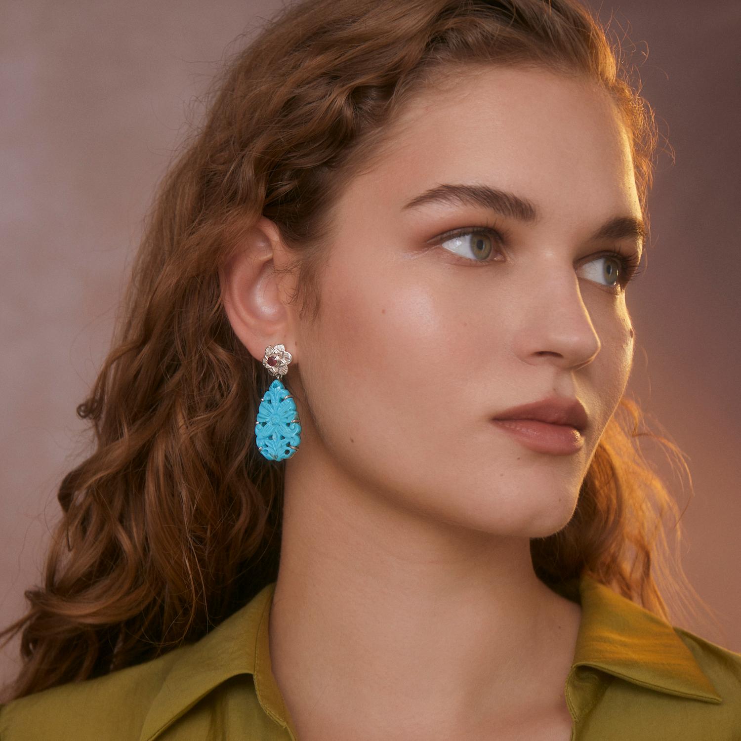 Ces magnifiques boucles d'oreilles en turquoise et rubis, uniques en leur genre, ont été fabriquées à la main dans nos ateliers. Elles sont composées de turquoises sculptées à la main et surmontées de rubis qui sont sertis dans des motifs floraux
