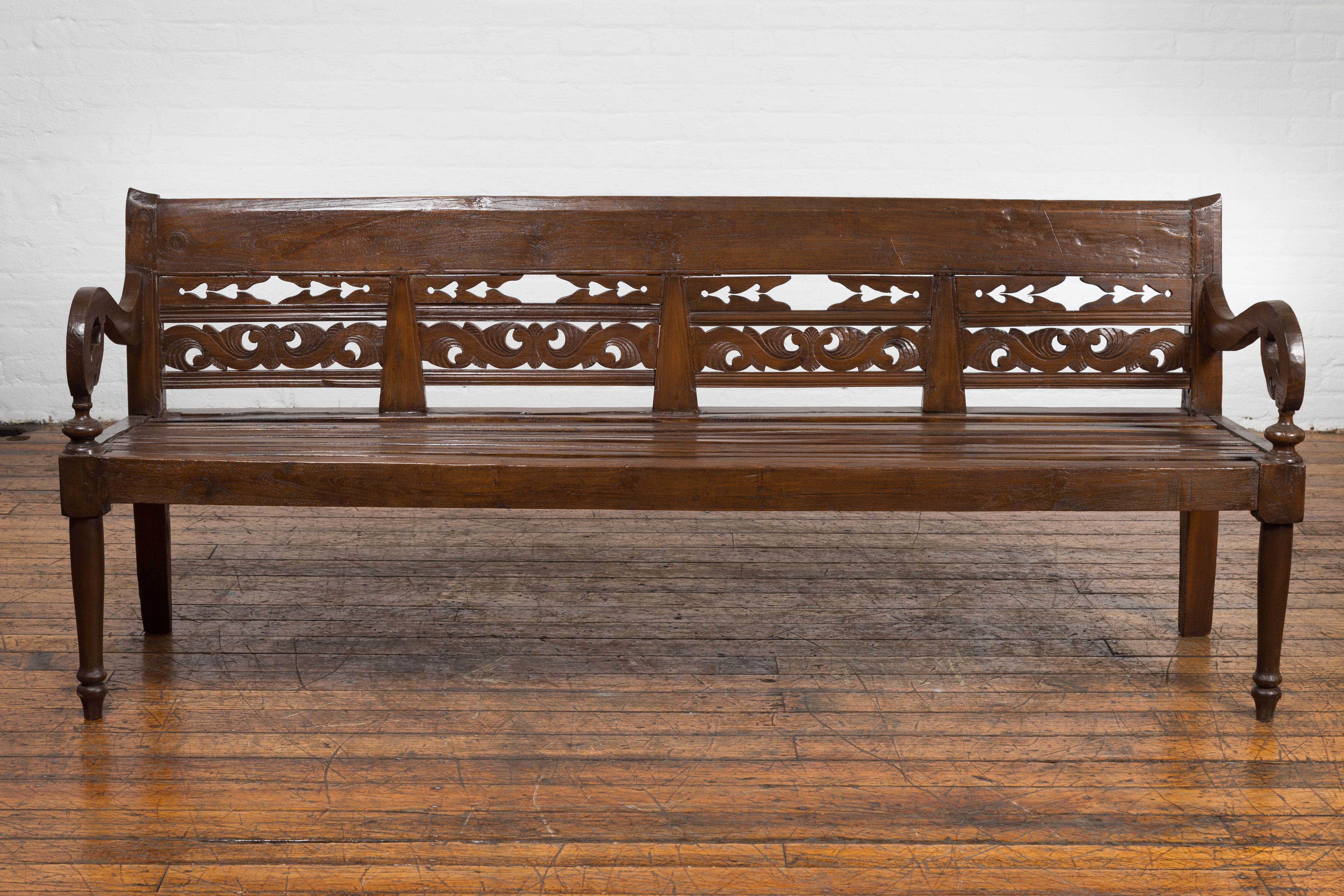 An antique teak wood settee from circa 1900 with hand-carved back, scrolling arms and turned legs. This Turn of the Century teak wood settee, dating back to circa 1900, is an exquisite blend of form and function. The backrest stands as a testament