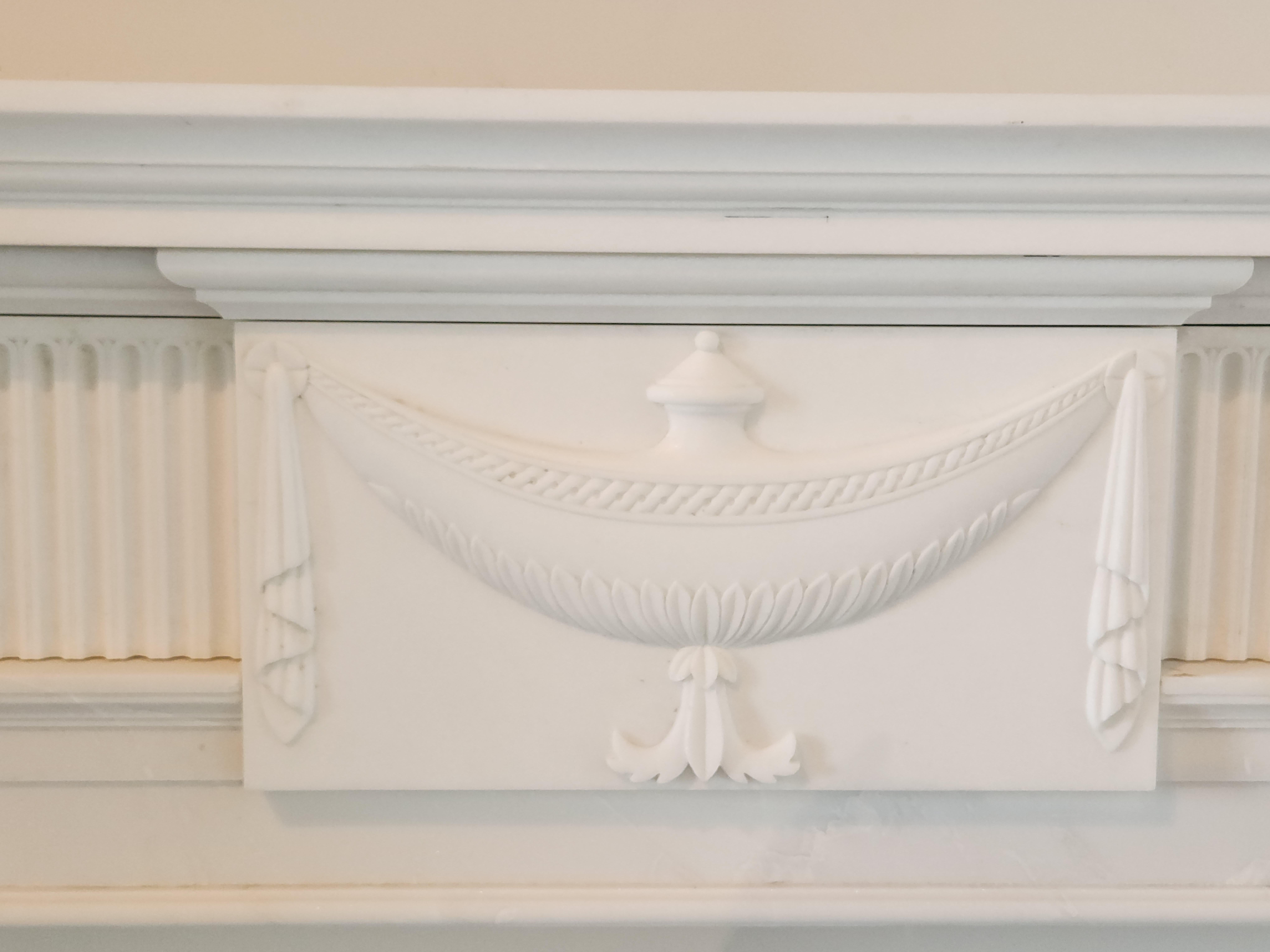 This beautiful Regency-style fireplace mantel is hand-carved in elegant white marble. The marble is a bright white, and is very clean, with very little veining. The hand-carved details on the fireplace include delicately-carved urns on either side