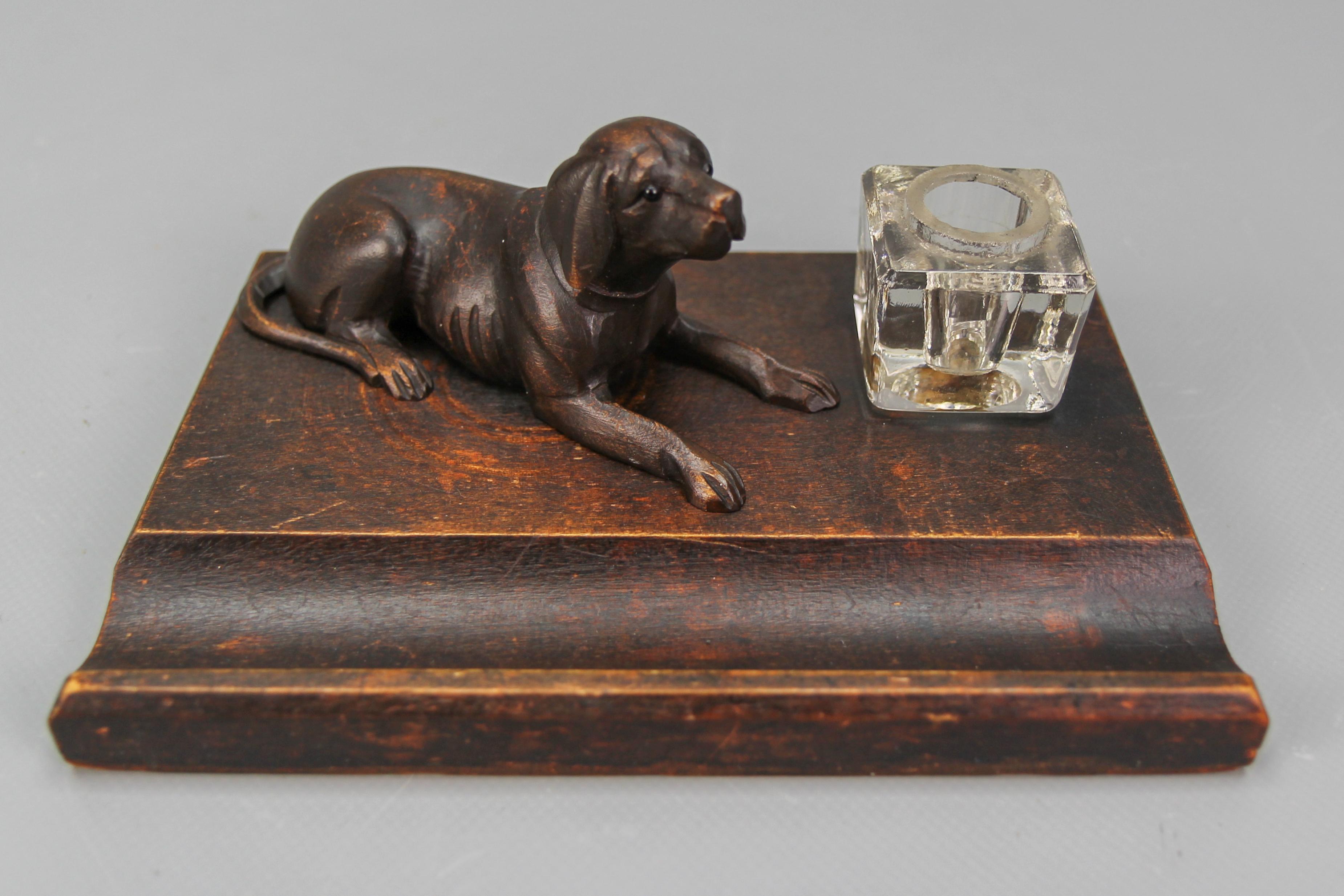 Beautiful Black Forest style hand-carved linden wood inkwell with a realistic dog figure with glass eyes. The stand includes a designated rest for pens and accessories in the front, along with a glass inkwell pot that can serve as storage for