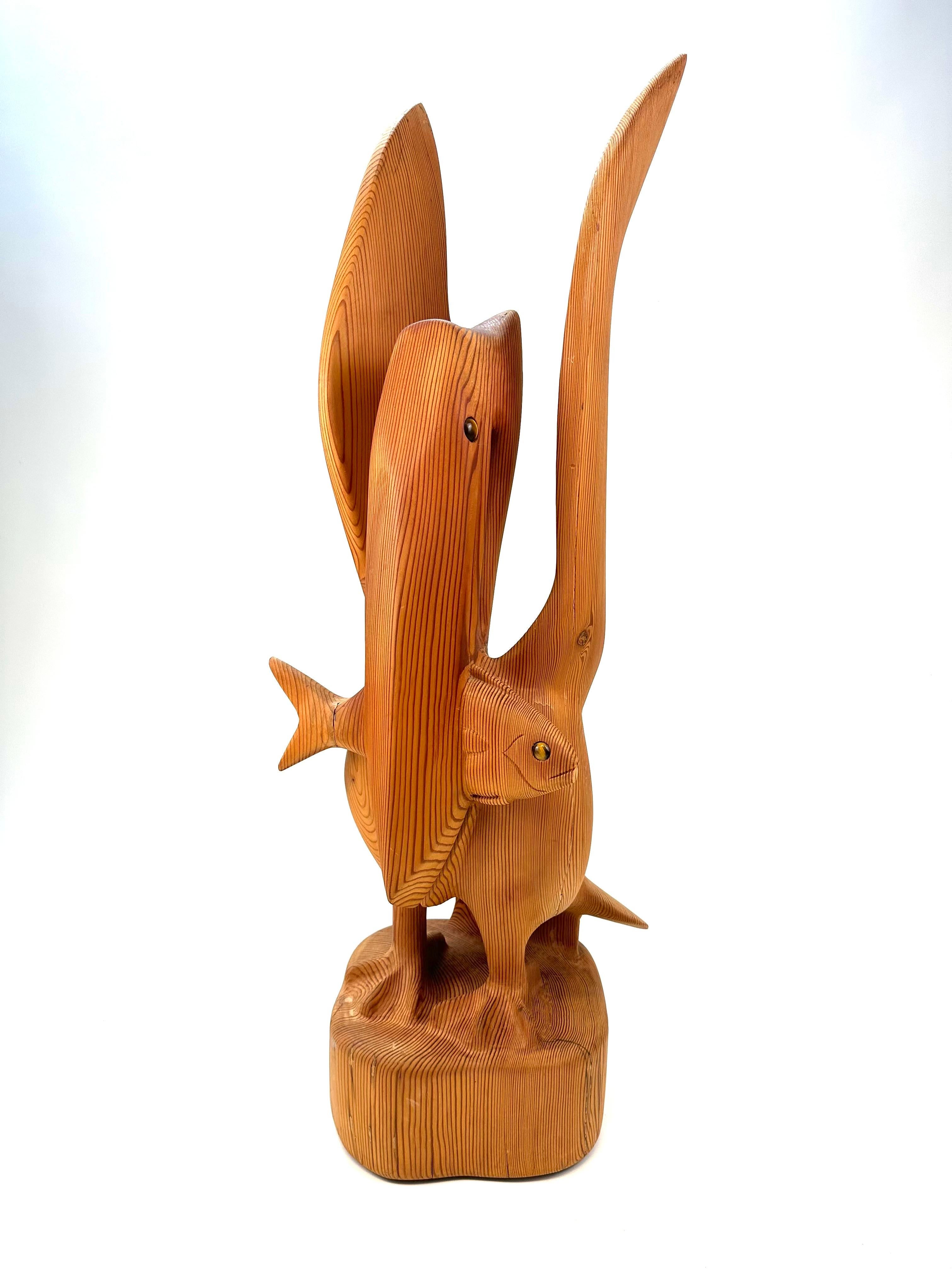 Una llamativa y caprichosa escultura de madera tallada a mano con un pelícano estilizado que sostiene un pez en el pico. Esta pieza única de estilo moderno de mediados de siglo está fabricada con madera maciza de Douglas Fur, mostrando bellos
