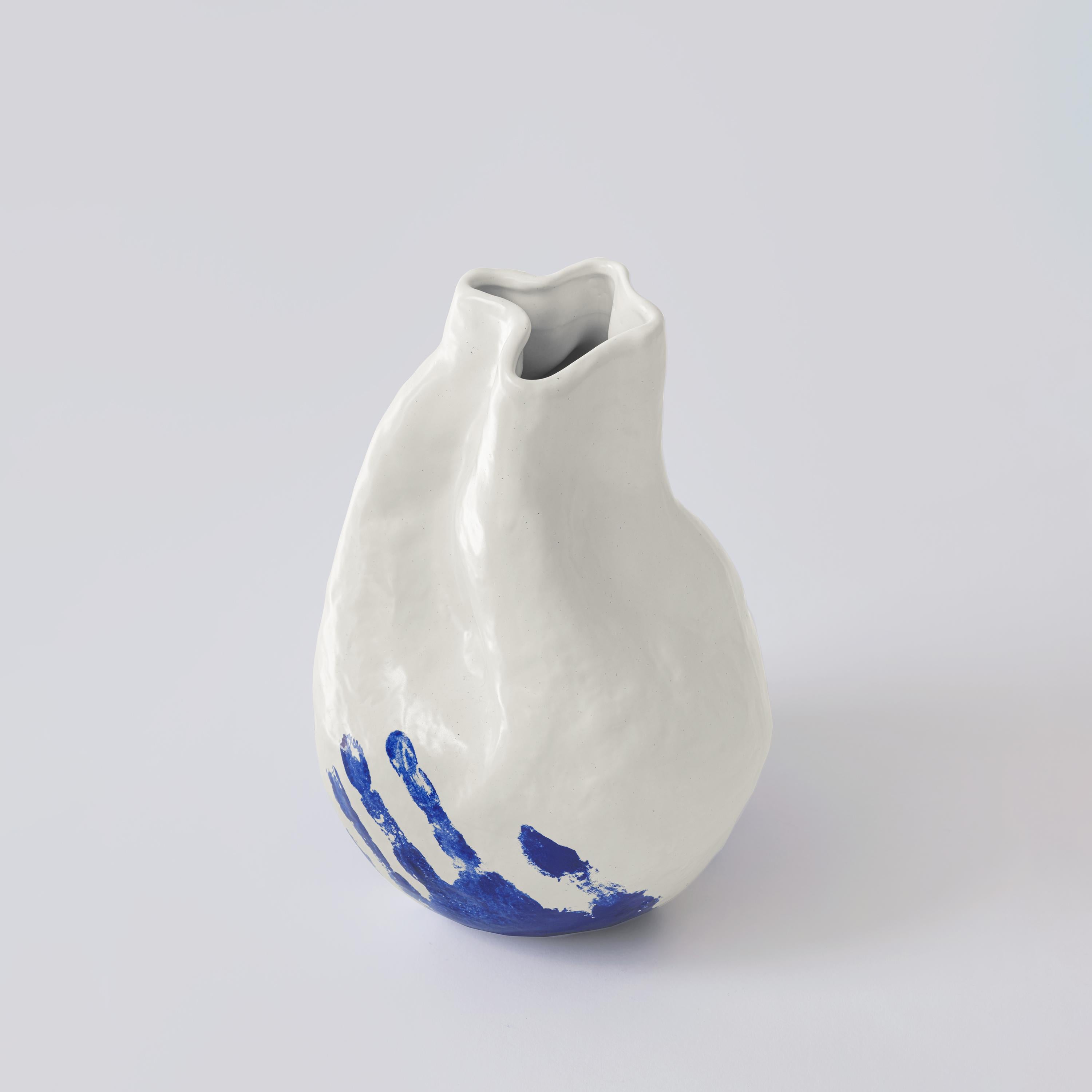 This porcelain hand-crafted vase from The Vibrant Touch Collection presents a tactile interplay of textures, with a glossy white finish that contrasts with a bold blue handprint at its base. The organic form of the vase, free from conventional