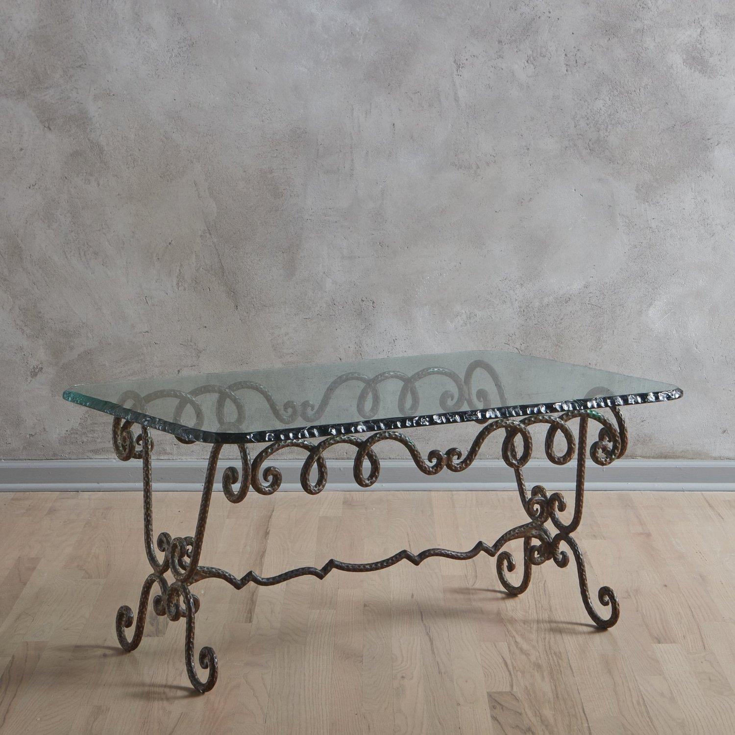 A 1960s Italian coffee table in the style of Garouste & Bonetti. This table features a sculptural, hand forged iron base with scroll details, a hammered finish and a center stretcher. It has a 1” thick rectangular glass tabletop with curved corners