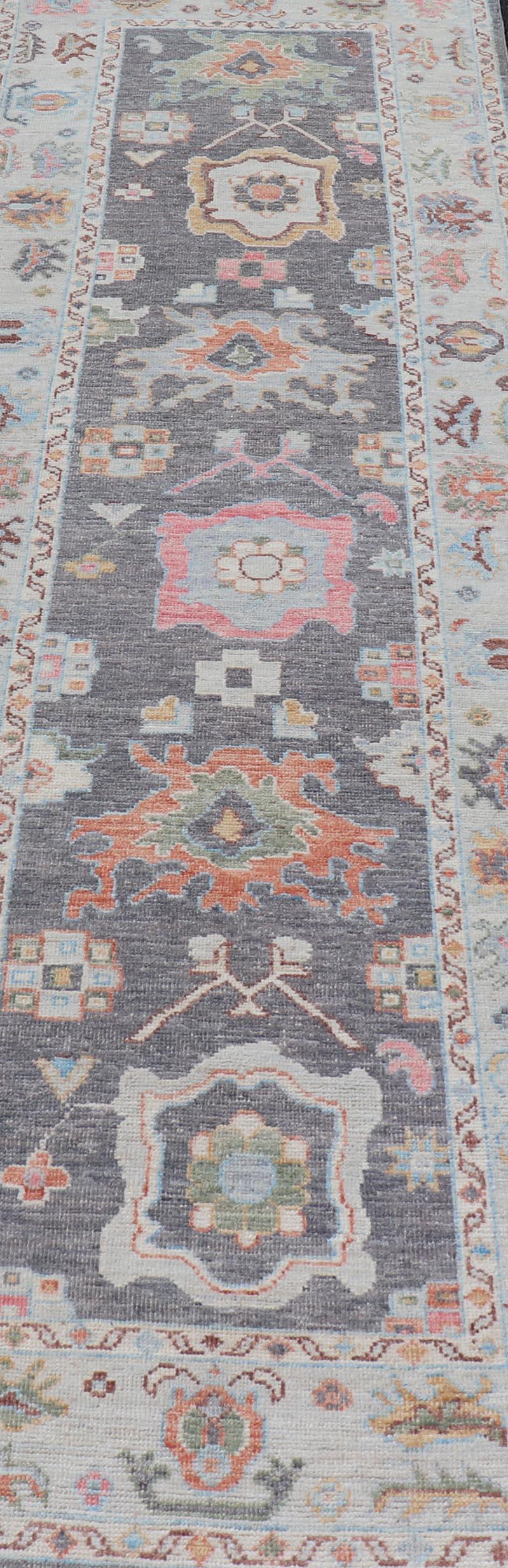 This Afghan Oushak runner features a grand motif, showcasing from the border to the field with a vibrant elegance that pops against the light blue-gray border, as well as the charcoal gray background throughout the center. The intricate details in
