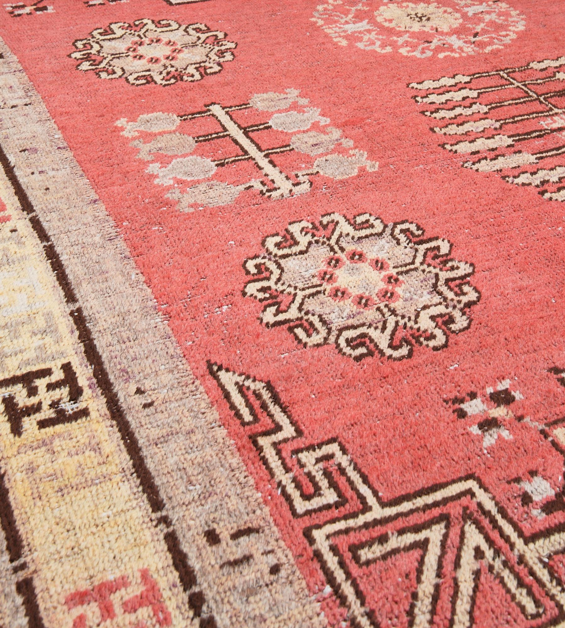 This antique, circa 1880, Khotan rug has a coral-red field with a central soft aubergine-purple square containing a soft yellow panel with a central rosette issuing floral motifs surrounded by a narrow coral-red band of linked circles surrounded by
