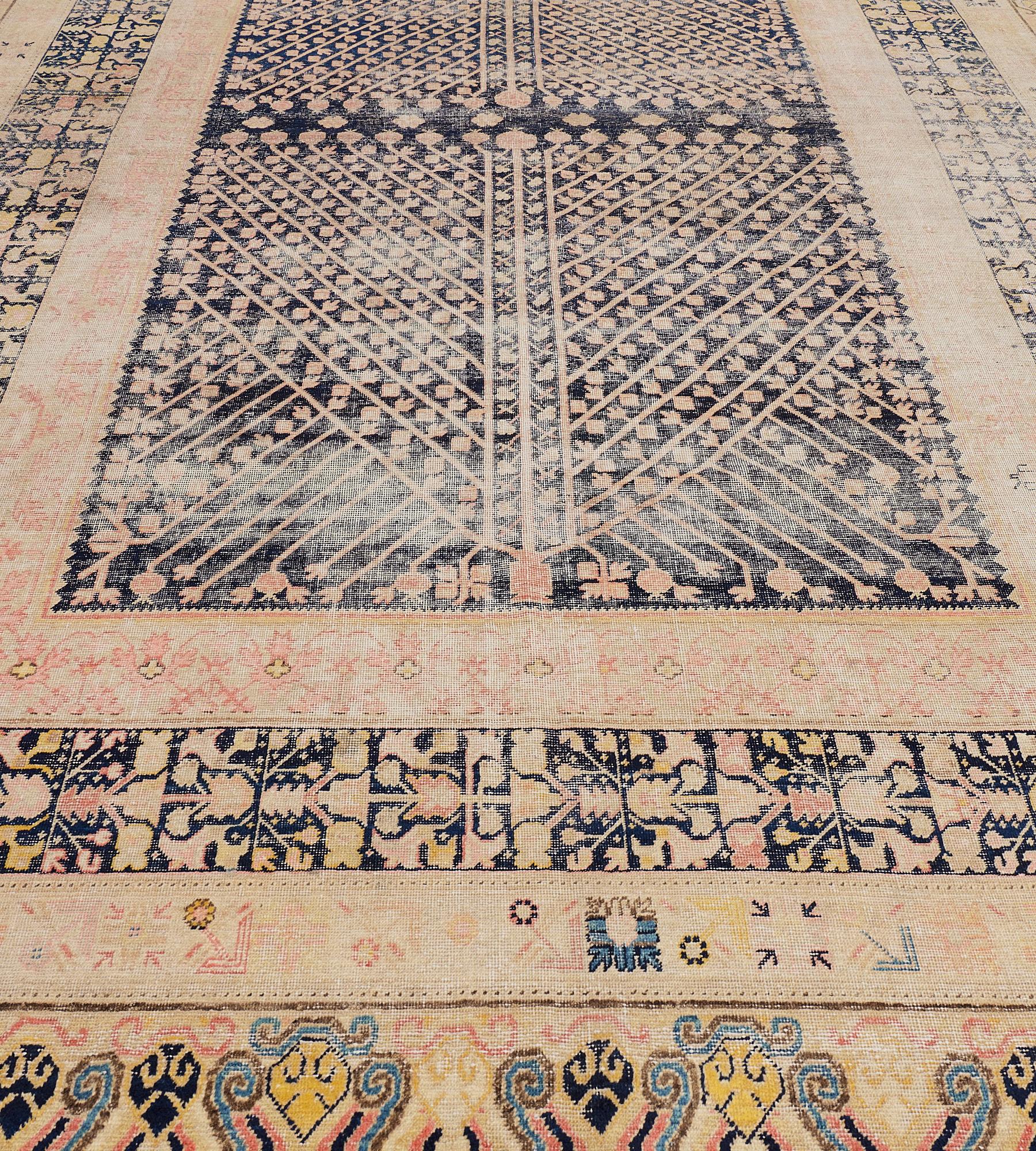 This antique, circa 1880, Khotan rug has a charcoal-blue field with two large panels of dusty-pink angular flowerhead and pomegranate vine, in a charcoal-black border of golden-yellow and shaded dusty-pink angular floral paneled vine between