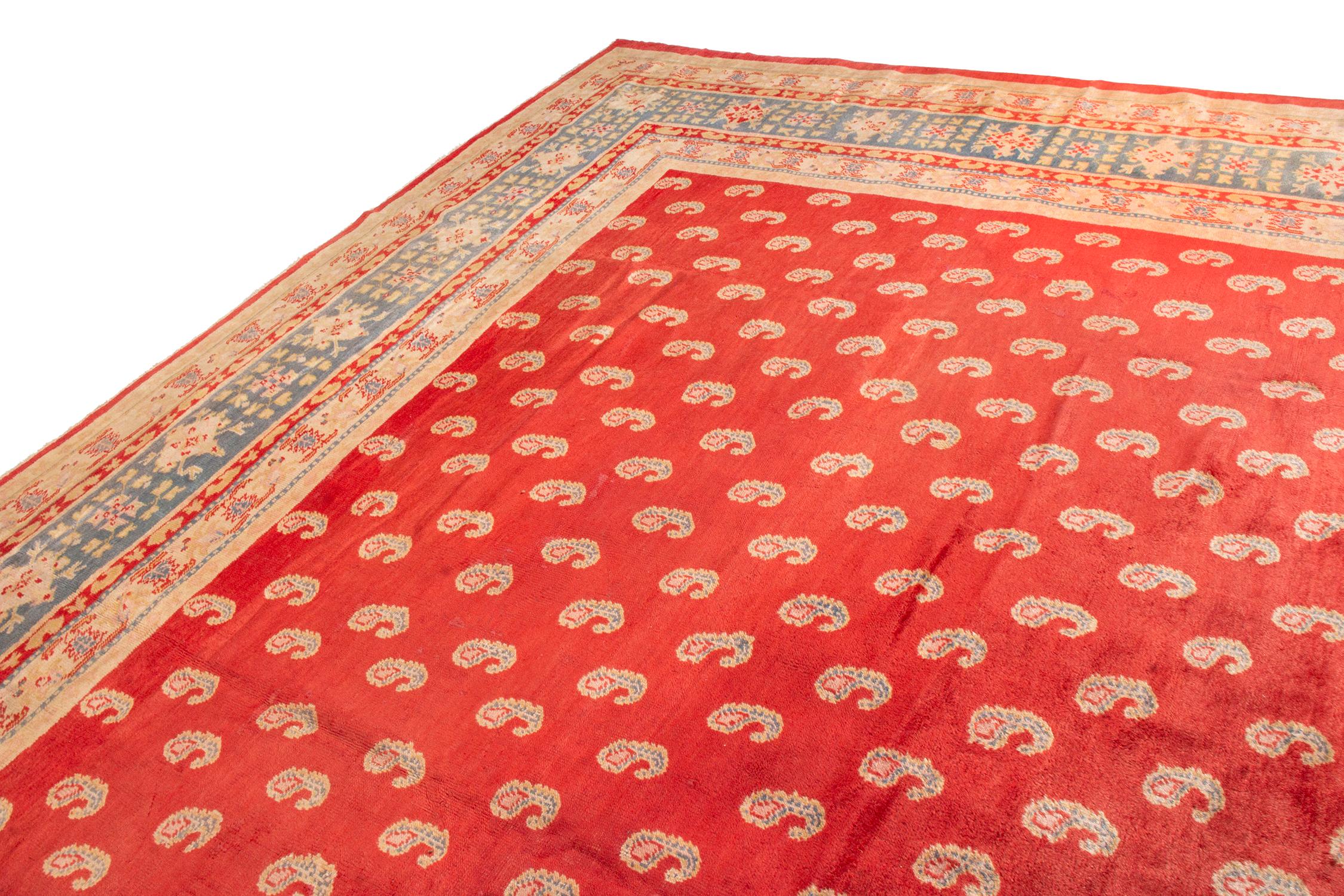 Hand knotted in wool originating from Turkey circa 1900-1910, this antique rug connotes a traditional Oushak rug design in rich red background, uplifting a venerated paisley pattern repeating in the same complementary beige-brown and blue hues