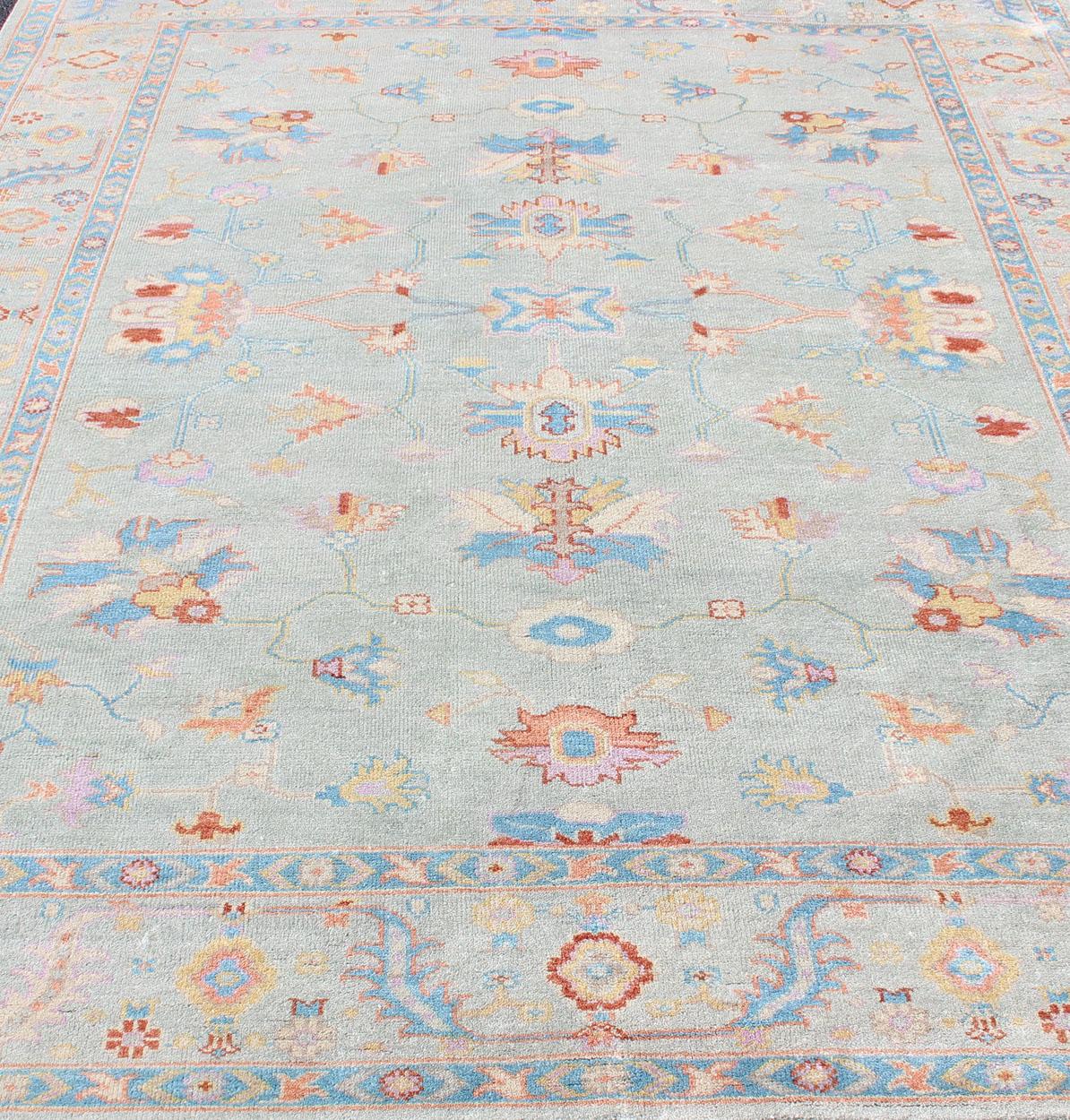 This wool hand-knotted Oushak rug by Keivan Woven Arts, was made in the 2010's. The field and border both have a cool blue gray. The field and border also share floral and leaflet motifs rendered in light blue, yellow, orange, blue, with small