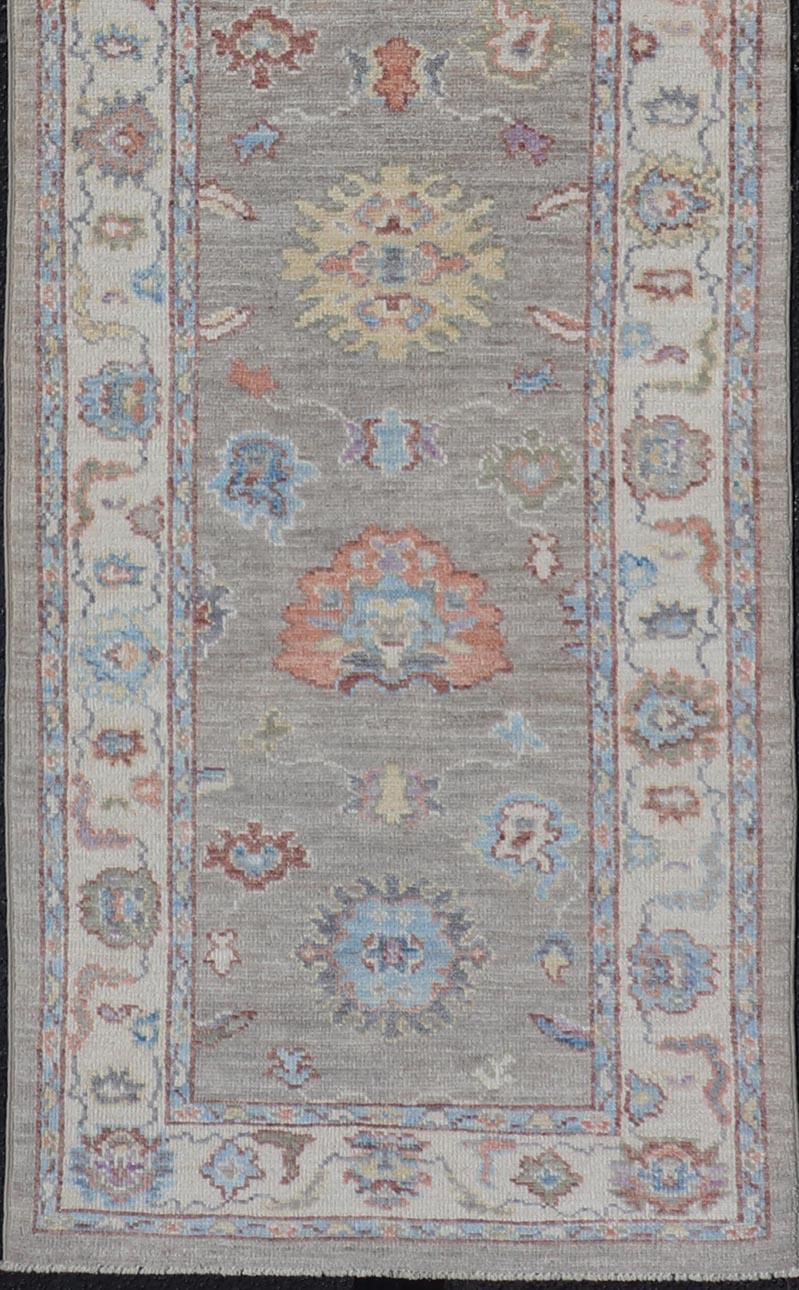This Oushak runner has been hand-knotted. The arabesque motifs scatter atop a light gray background. Within the field, bright displays of gold, teal, blue, pink, maroon, and hints of green sit above a classic gray background. These vivid colors
