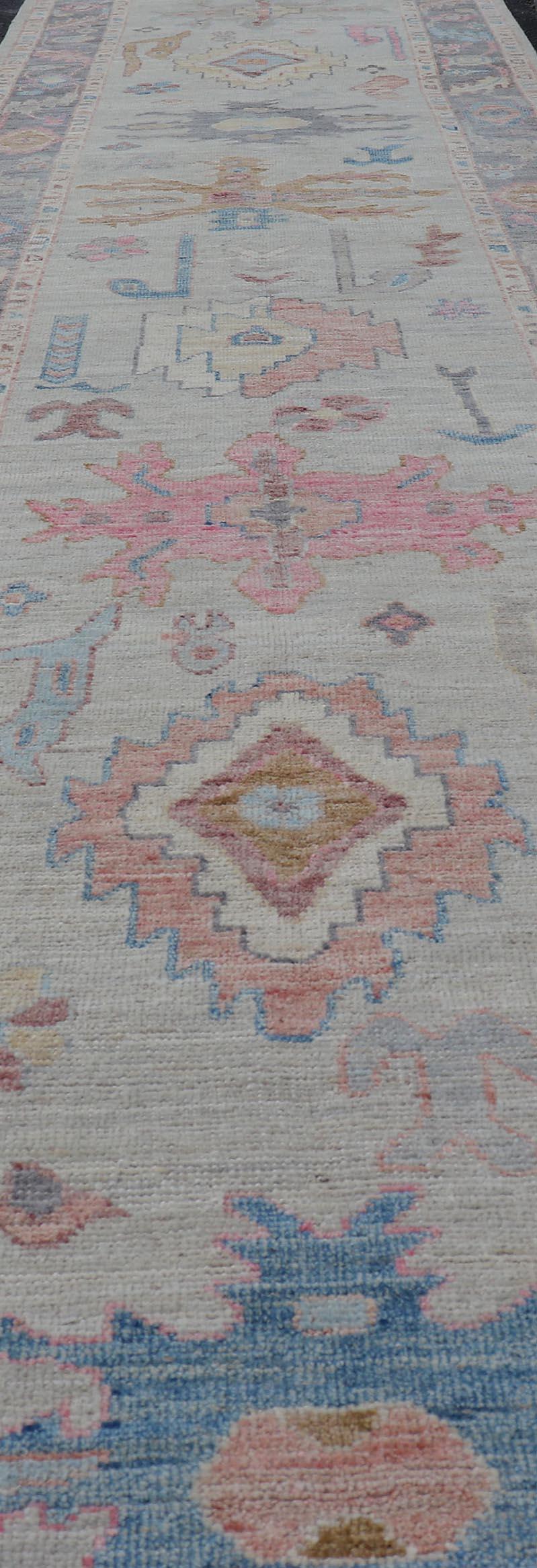 This long Oushak runner has a cream background with a light charcoal border. The border has a leaflet-like design is rendered in green, coral, and blue with a hint of yellow, while the main field's tribal design is rendered in a sandy bronze, gold,