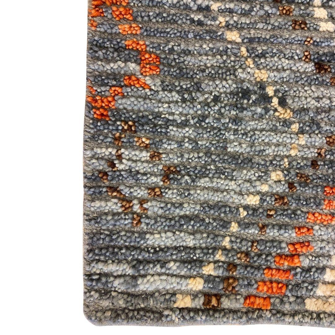 Questo tappeto tribale in lana annodato a mano nei colori grigio, blu, arancione e marrone, di dimensioni pari a 2' x 2', presenta una sorprendente combinazione di vello alto e basso che crea una superficie dinamica e strutturata. I vivaci accenti
