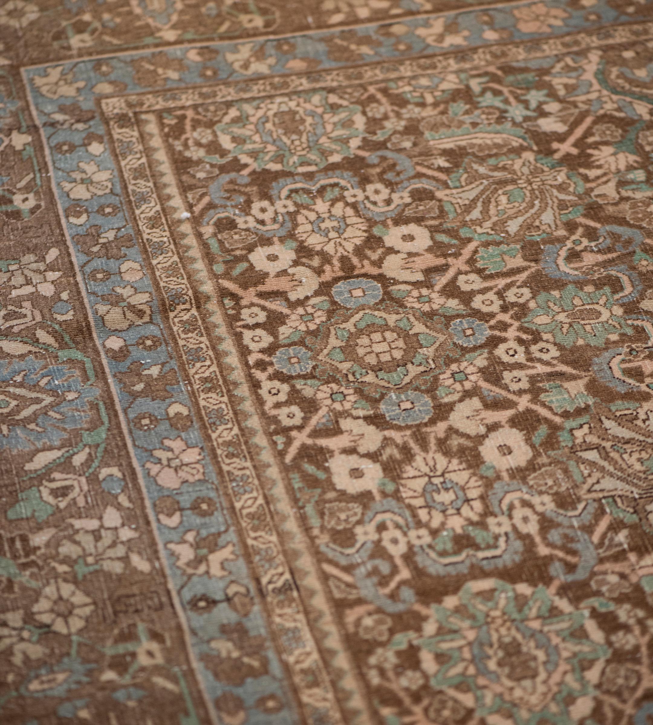 This antique, circa 1920, Persian Tabriz rug has a soft brown field with an overall pattern of light blue, bottle-green, ivory and sandy-yellow bold palmettes surrounded by similarly colored scrolling leafy vine linking flowerheads, in a soft brown