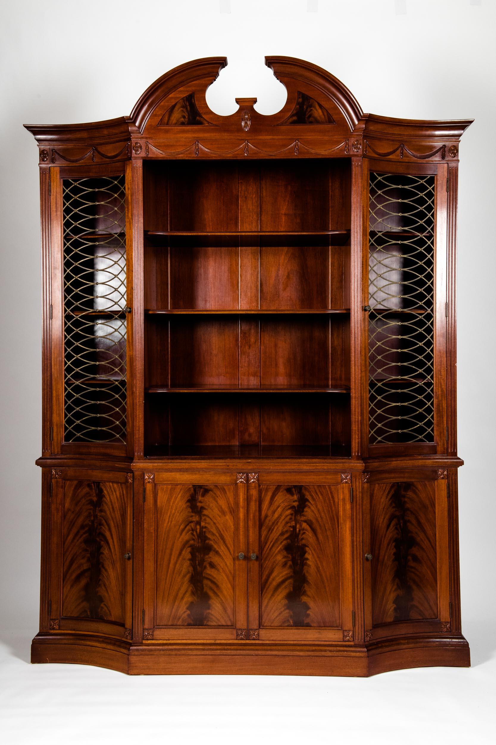 Handmade 20th century mahogany wood breakfront or cabinet with arch top and carved design details. Two parts, upper section with pediment top over swag carved panel with four shelves flanked by grill work side doors. Stand on base with four doors.