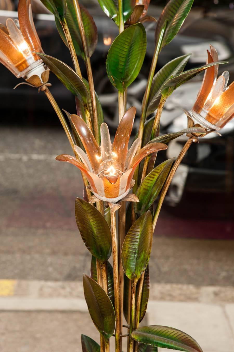 Exotic standard lamp of painted metal stalks with leaves supporting four hand blown glass flowers in delicate shades of umber, standing on a twisting circular base.