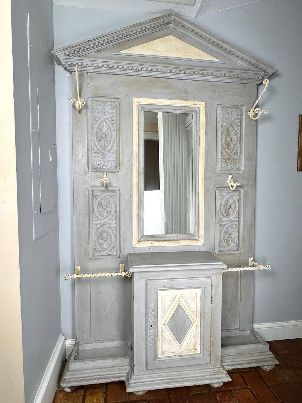 Albero da sala dipinto e intagliato a mano con specchio e ganci. Un pezzo perfetto per la stanza del fango estiva e balneare  per appendere borse, cappelli e ombrelli. Un'anta dell'armadio si apre su un ripiano per riporre i guinzagli del cane o i