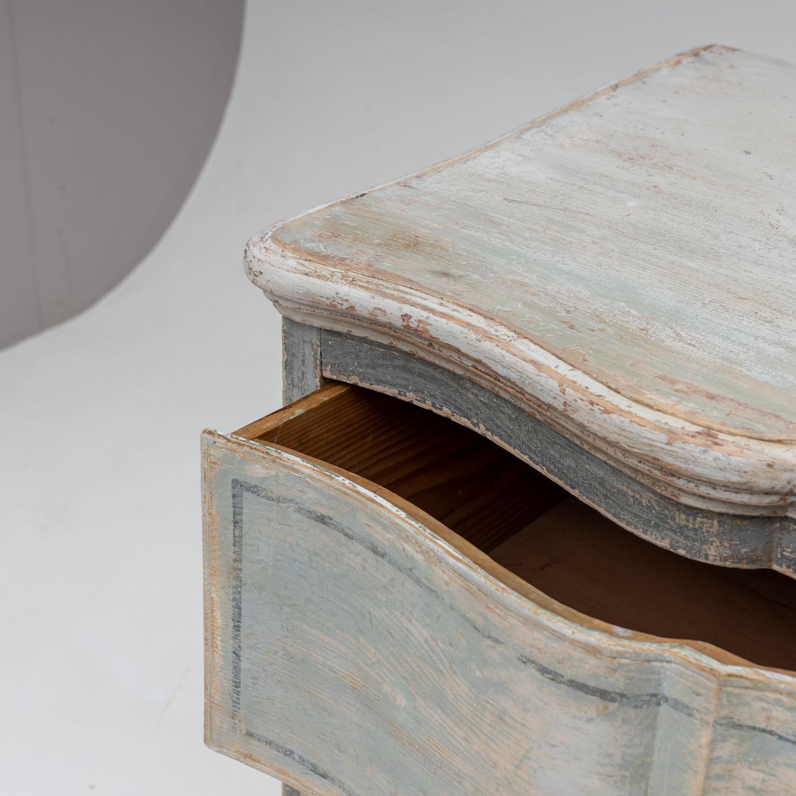 Two-drawer baroque-style chest of drawers standing on a cut-out base. The body with a slightly wavy front has been repainted blue-grey according to historical models and decoratively rubbed through.