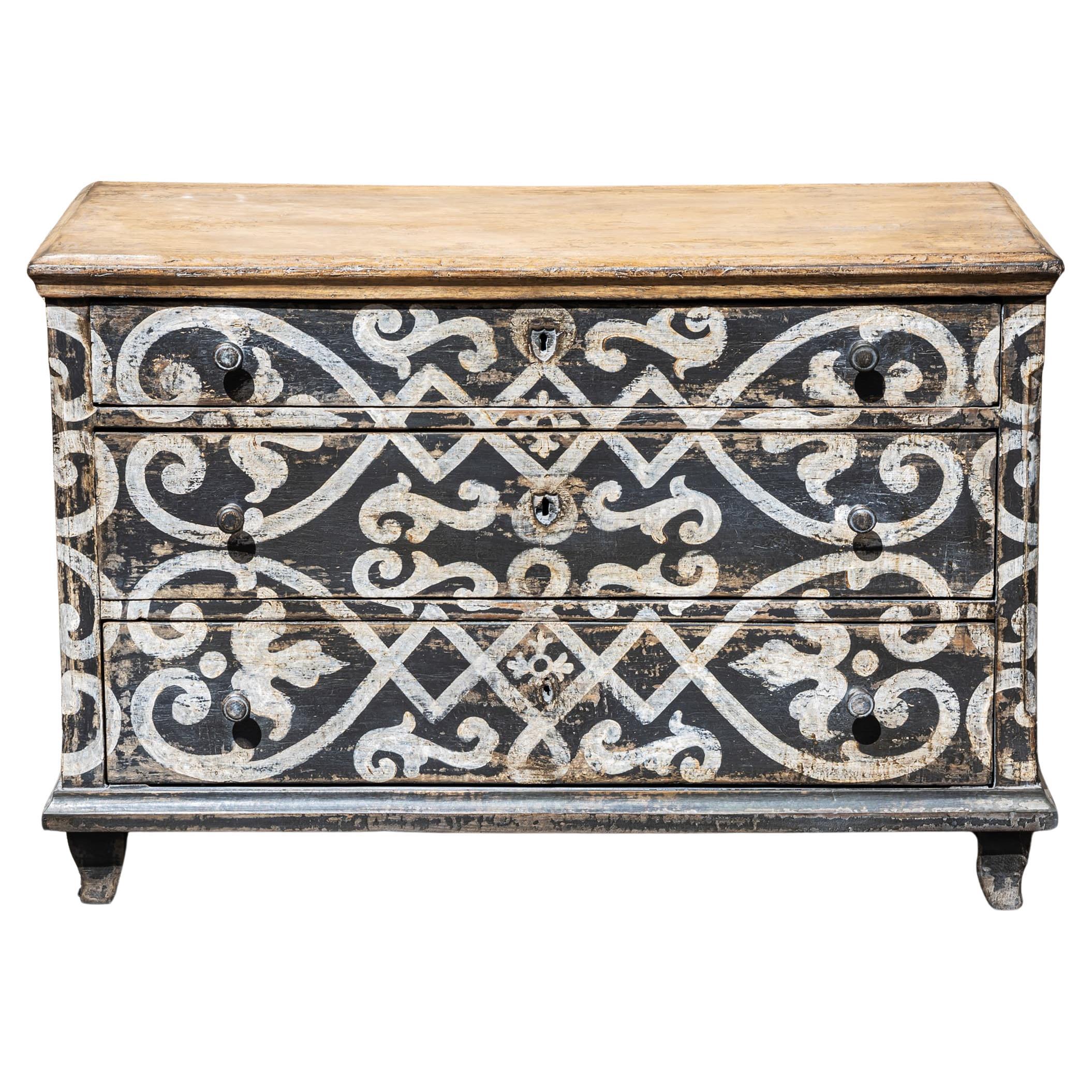 Hand-painted three-drawer commode with a dark gray base and cream-white decoration featuring hearts and ribbons, inspired by historical designs. The corners are slightly chamfered, and the top is finished in a light brown tone. The surface has been