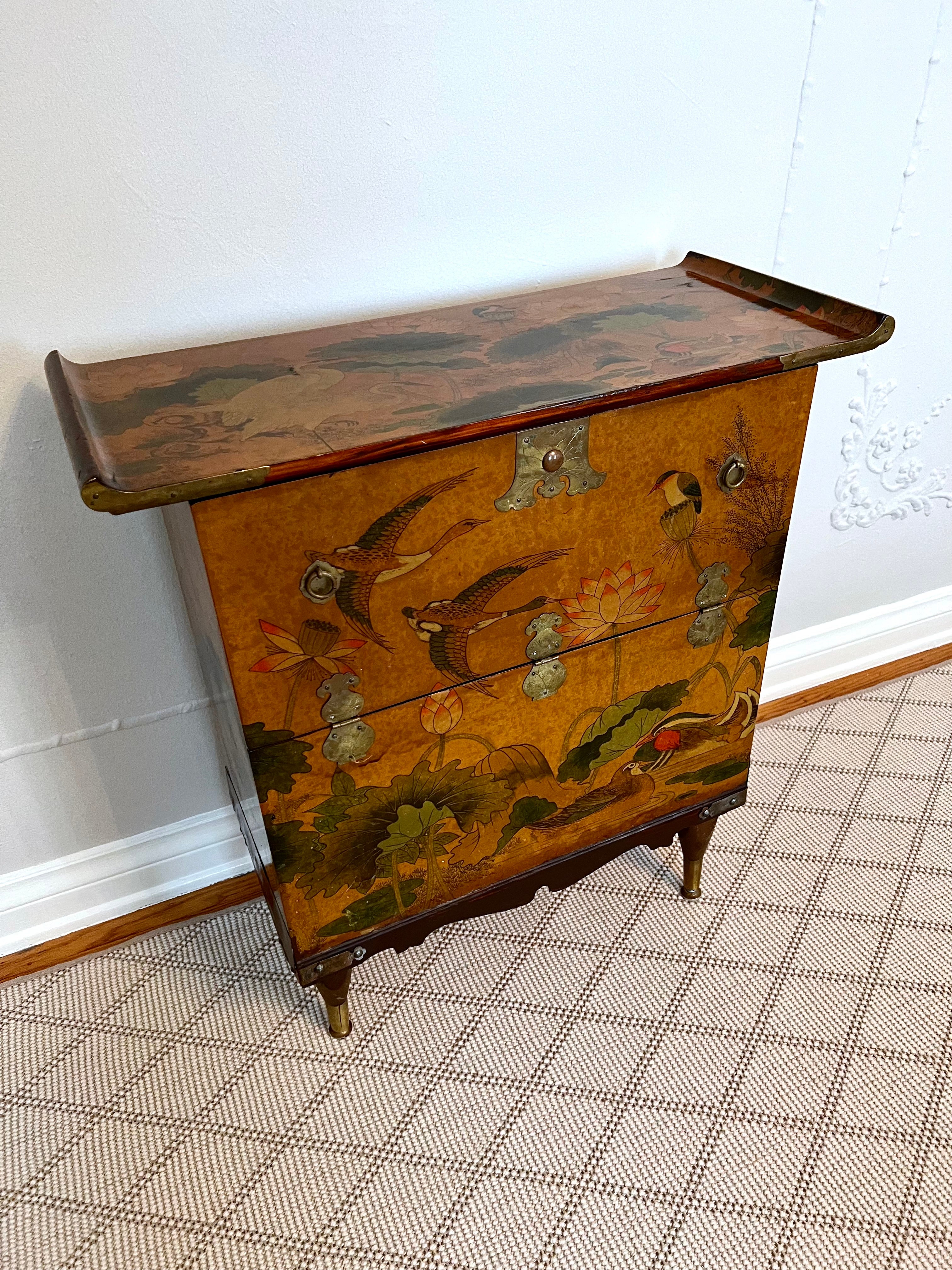 Beautiful Asian cabinet featuring upturned corners and Chinese script lining the interior. The top and front surfaces feature hand-pained lotus and bird detail for a natural scene. 

This cabinet can be used to store blankets or tall items as it