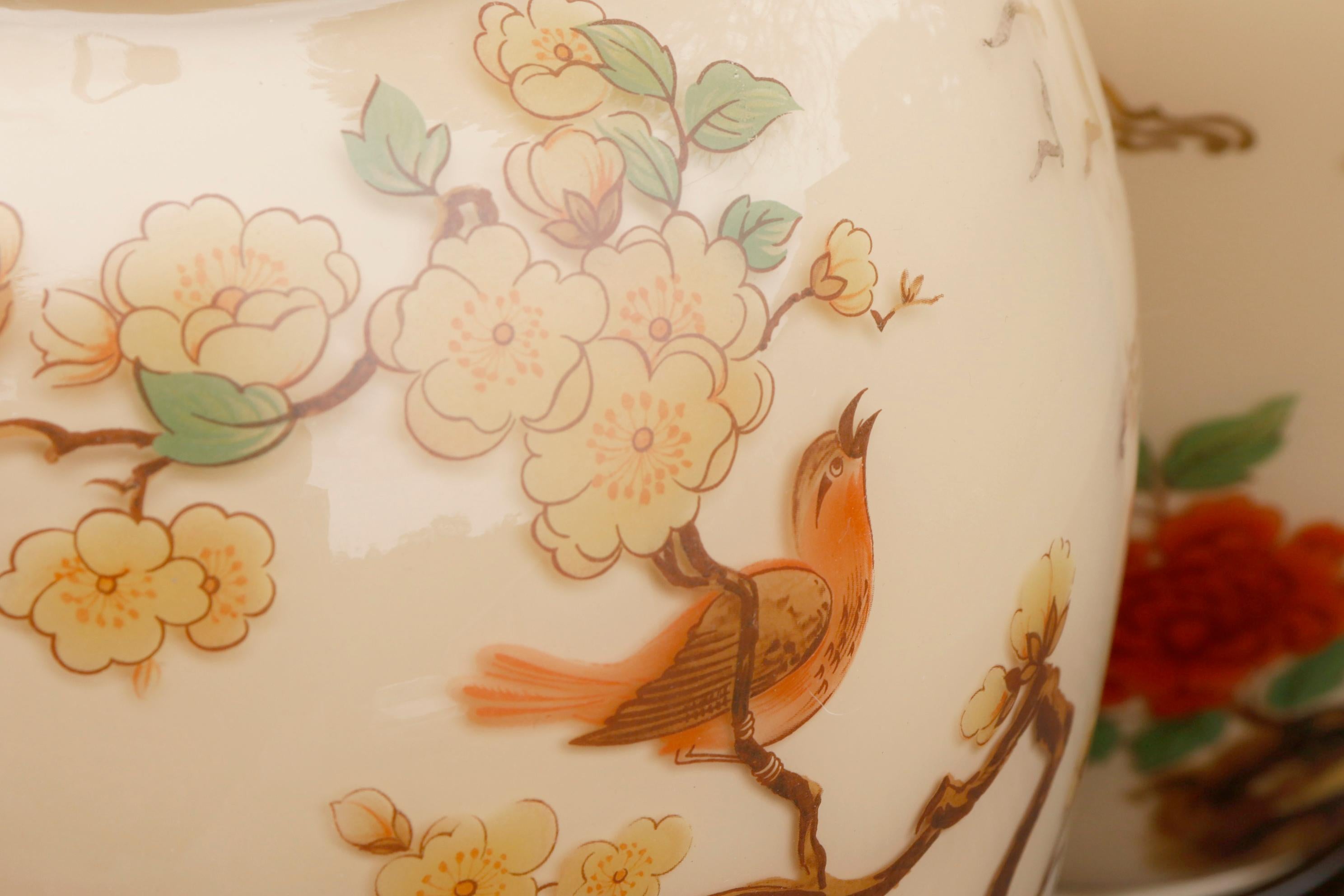 A pair of hand painted Chinoiserie style glass table lamps. Decorated on the outside with an Orange Headed Thrush resting on a cherry blossom branch and an Asian Brown Flycatcher nestled amongst roses. Painted on the inside in a light taupe. Stands