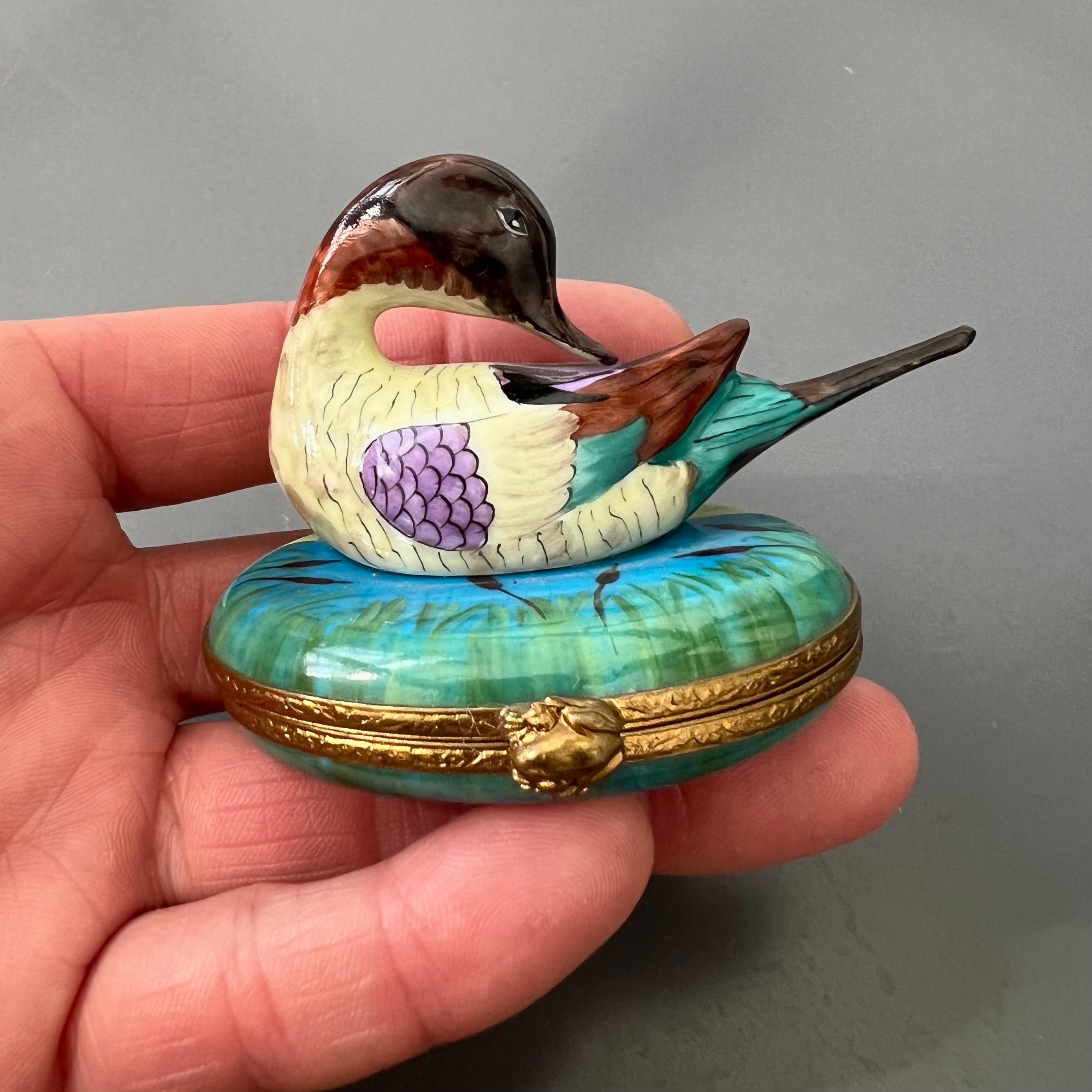Ce charmant coffret à bibelots en porcelaine de Limoges représente un canard à queue fine magnifiquement peint à la main, reposant sur une base de nénuphars. Il présente des détails exquis et des couleurs vives. Le canard sculpté est peint dans des