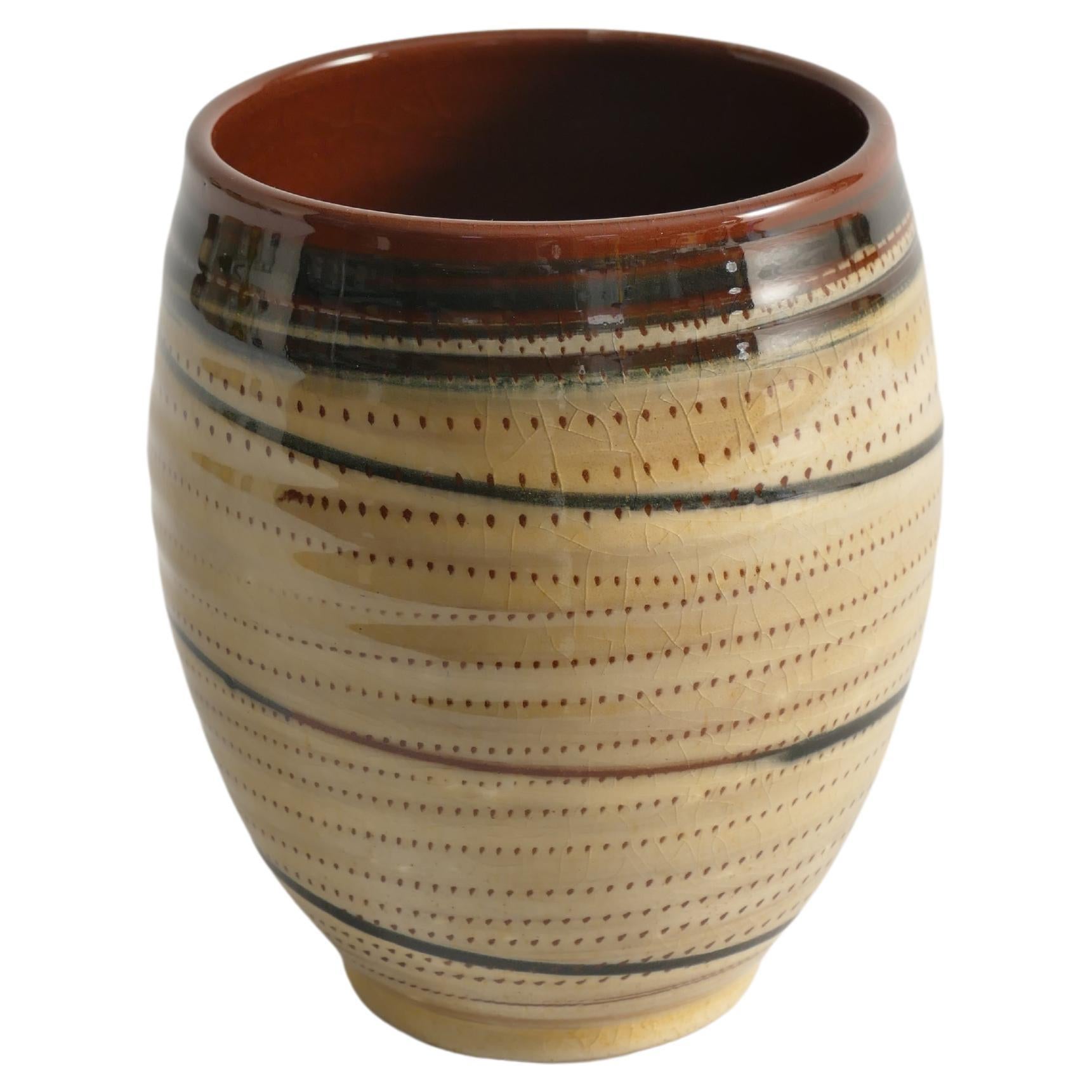 Ce vase suédois peint et tourné à la main présente une glaçure brillante de couleur crème ornée de motifs graphiques peints à la main dans des tons terreux. Le décor se compose de lignes pointillées croisées et de lignes entièrement connectées, ce