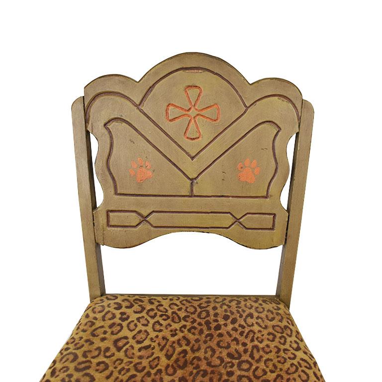 A whimsical hand painted wooden chair with upholstered leopard print cushion. The back of this piece is wood, with a stylized carved design in orange on a green background. The legs and sides are painted a muted green with orange details. The back