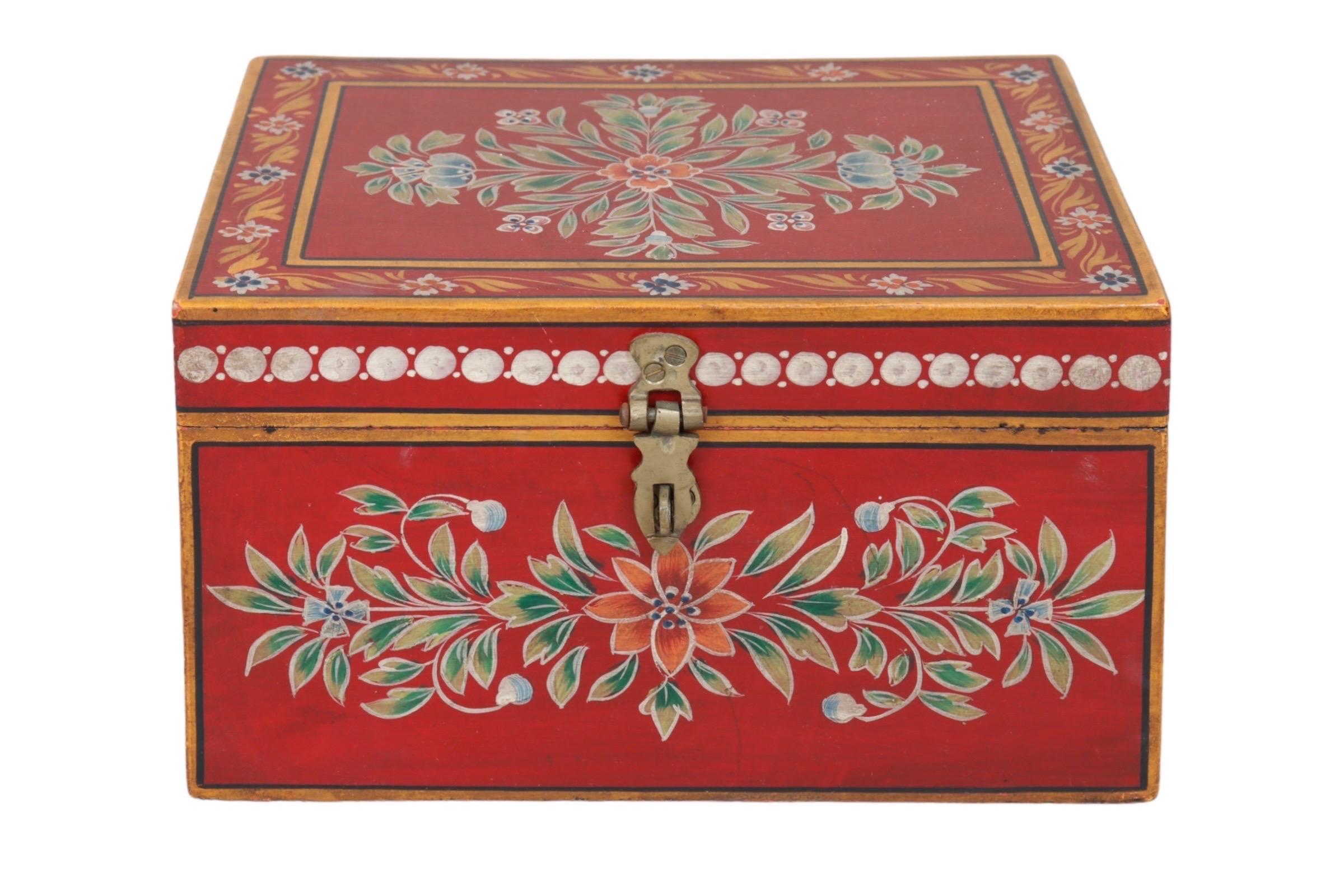 Una caja de baratijas de madera pintada a mano. Ricamente decorada con aerosoles florales en cada superficie, con flores naranjas y capullos azules a lo largo de follaje verde sobre un fondo rojo brillante. Bordes dorados perfilados en negro, con