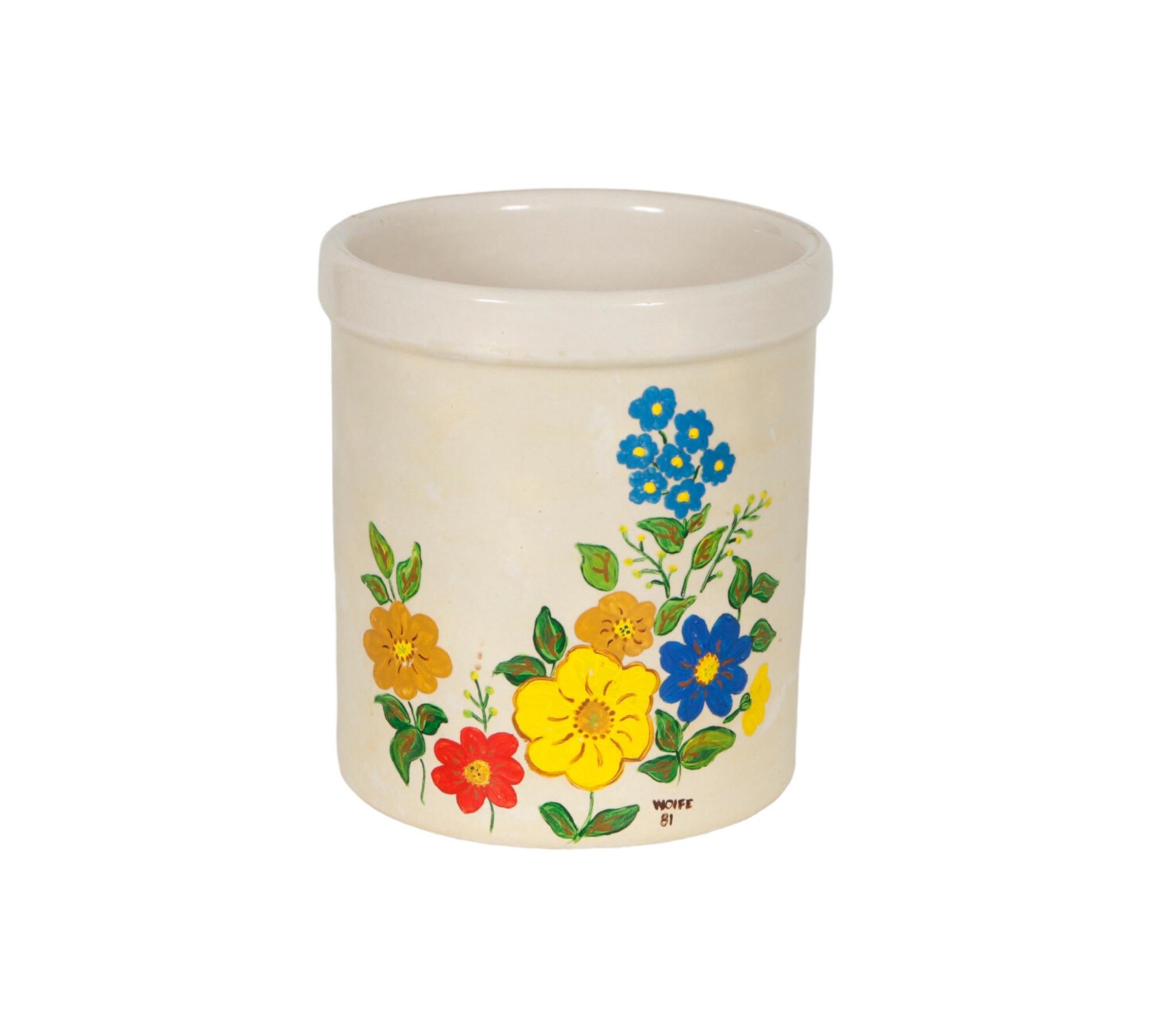 A hand-painted stoneware utensil holder jar. The front side is decorated with simple flowers in primary colors over a beige base. Signed Wolfe ‘81.

