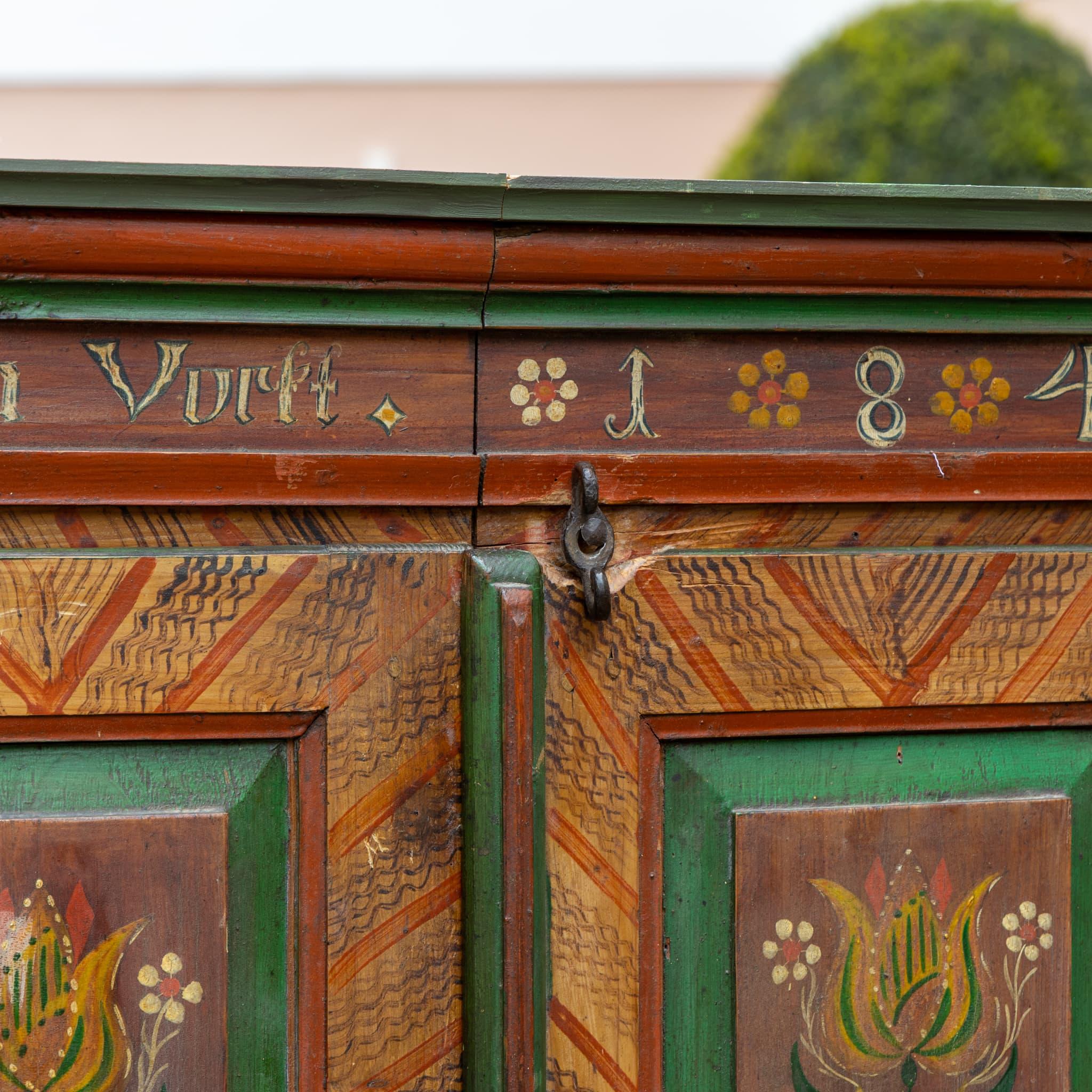 Armoire folk a due ante vivacemente decorato con motivi floreali simmetrici su quattro pannelli centrali, incorniciati in verde e ornati da disegni di tulipani e fiori. Motivi geometrici e puntiformi arricchiscono le superfici circostanti. Sul