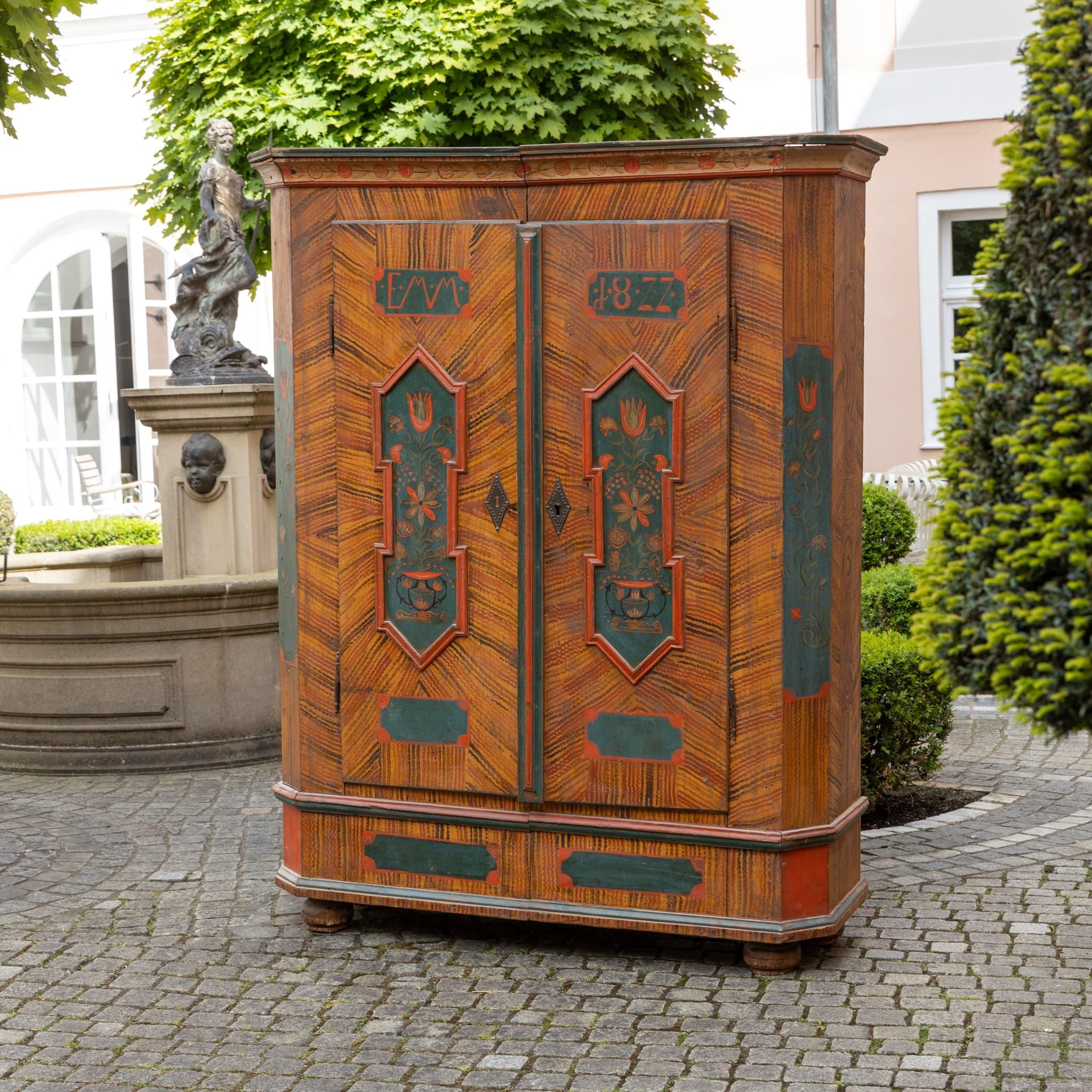 Armoire folk a due ante con vivaci dipinti floreali sui pannelli delle porte e sulle sezioni laterali. Sulle porte sono incisi il monogramma Emme e la data 1877. Il pezzo è caratterizzato da uno sfondo a grana grossa in tonalità calde, contrastato