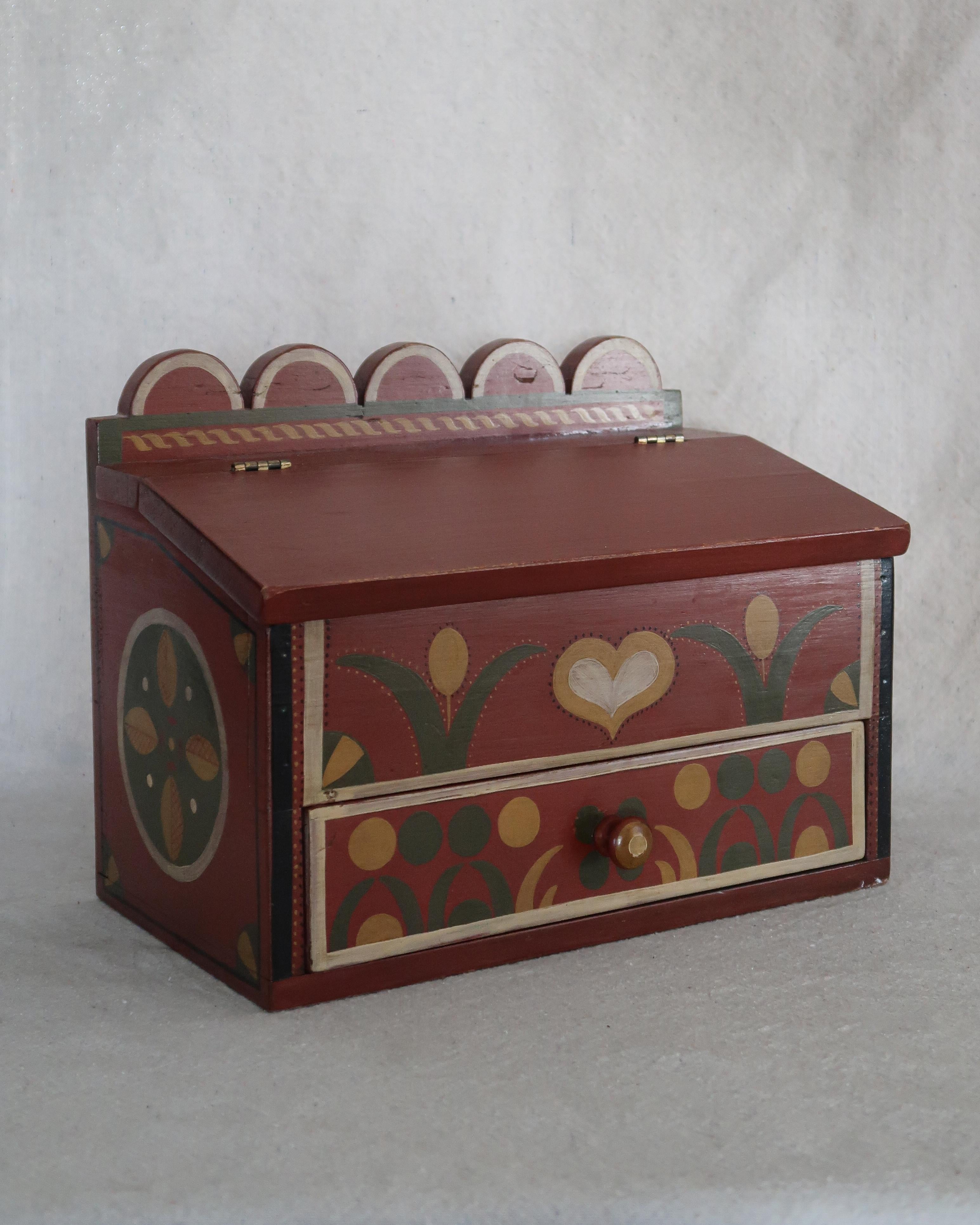 El folk moderno se une al encanto de la vieja escuela. Esta caja de estilo rústico pintada a mano tiene una tapa inclinada con bisagras, un cajón inferior y todos los dulces detalles -corazones, tulipanes, festones- que hacen un guiño al diseño