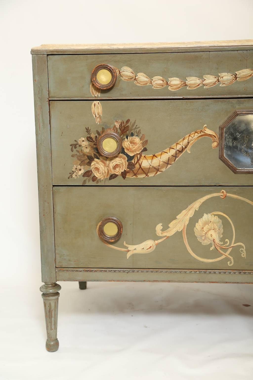 Commode having a molded top of faux marble its case hand-painted with classical motifs of cornucopias scrollwork and husking a central, etched églomisé plaque decorates it's middle drawer with round handles inset with a mirrored disk raised on round