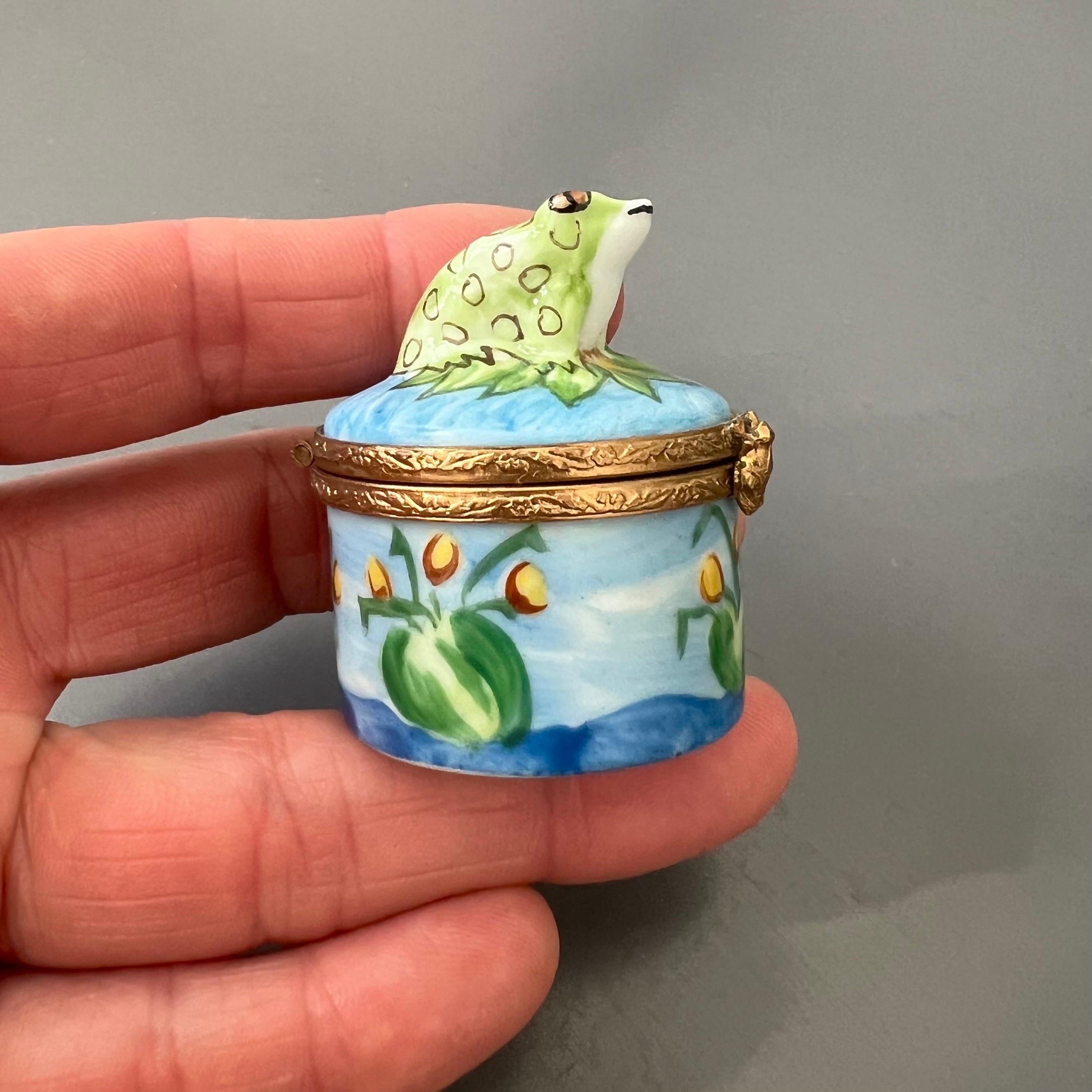 Délicieusement fantaisiste et méticuleusement réalisée, cette boîte à bibelots en porcelaine de Limoges présente une scène pastorale peinte à la main avec une charmante grenouille verte perchée au sommet d'un nénuphar. Le couvercle s'ouvre pour