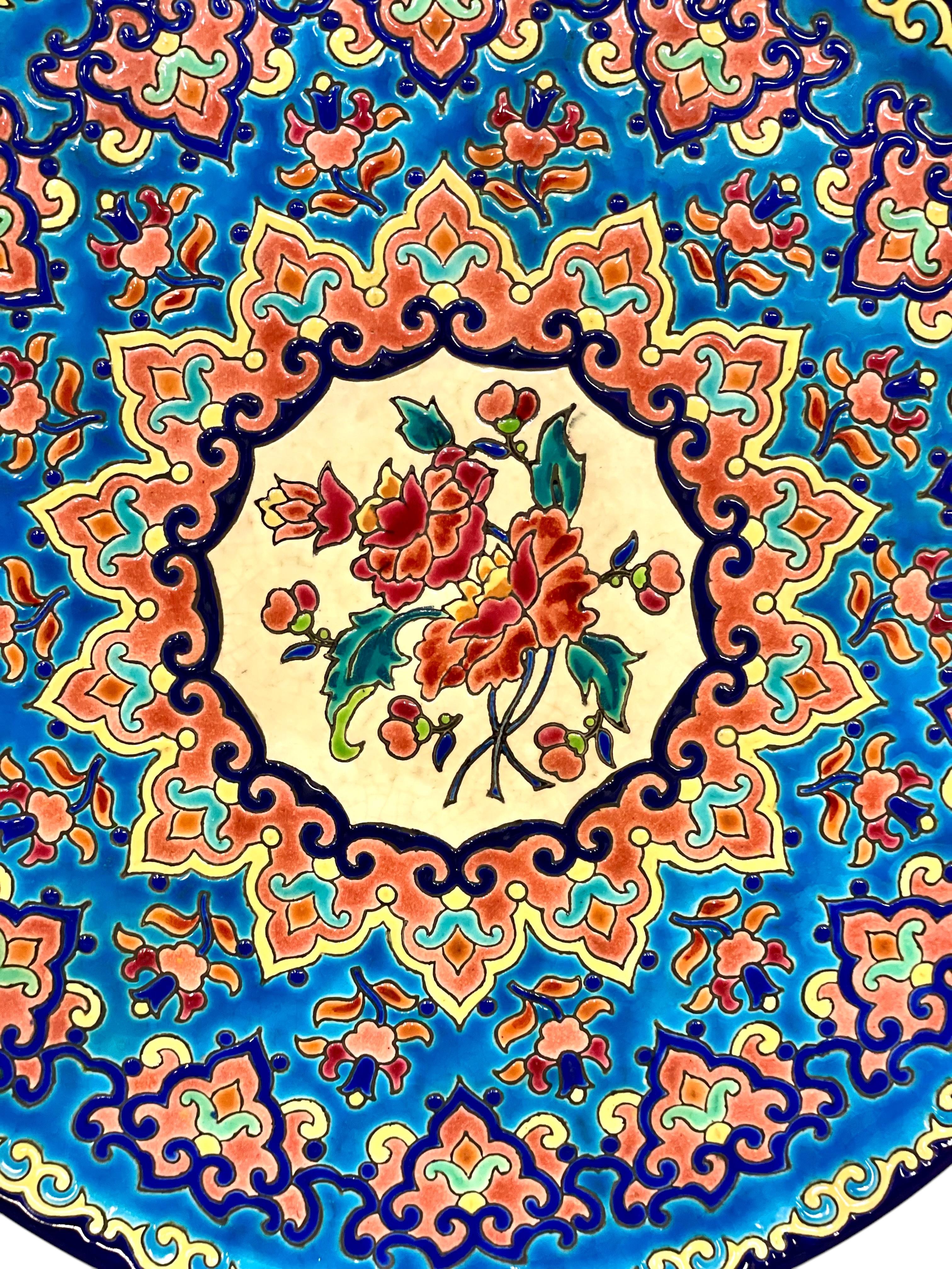 Questo squisito centrotavola è un vivace esempio delle rinomate ceramiche smaltate Longwy, realizzate a mano in Francia e riccamente decorate con intricati motivi floreali e geometrici policromi. Al centro di un bouquet di peonie in fiore, il piatto
