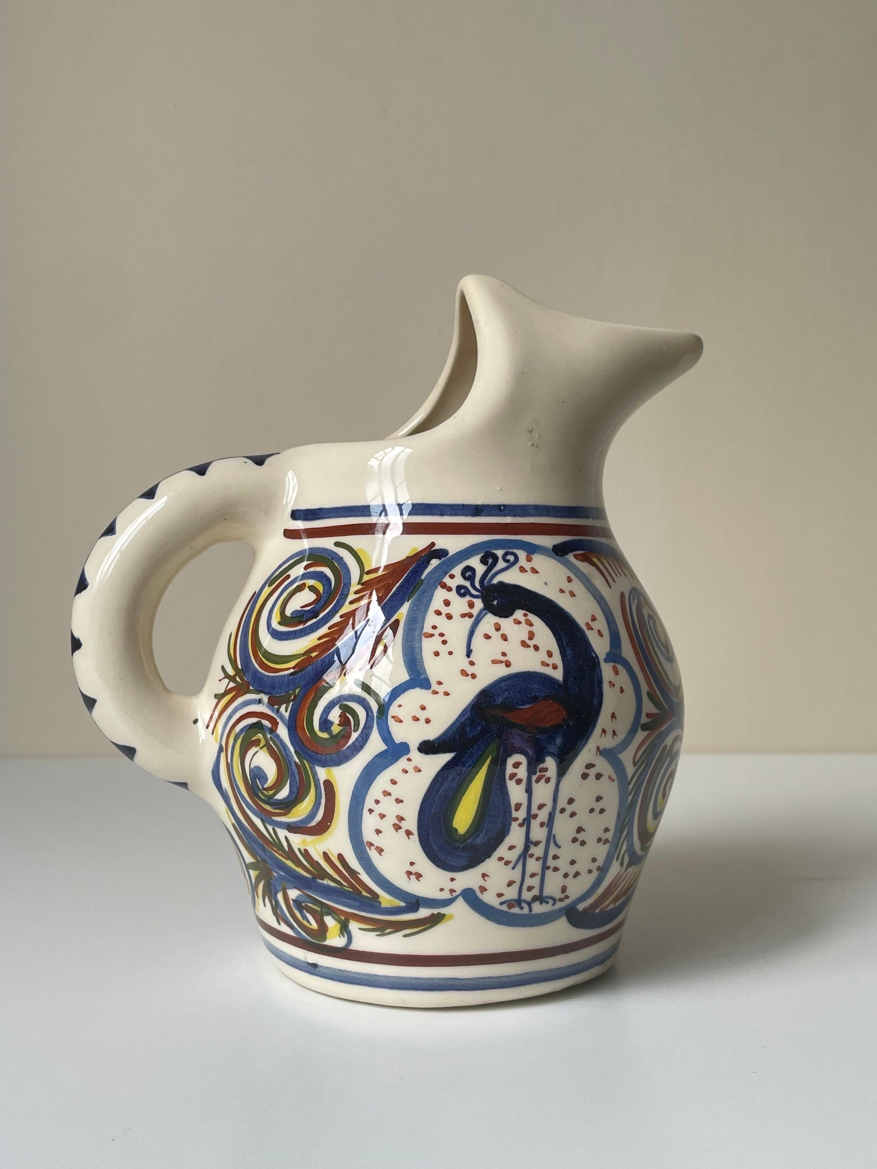 Multicolored hand-painted ceramic pitcher vase. Decorated with multiple lined, swirling, triangular, dotted and organic motifs with a stylized peacock bird in the centre on each side. Blue, brown and yellow colors under clear glaze. Signed under