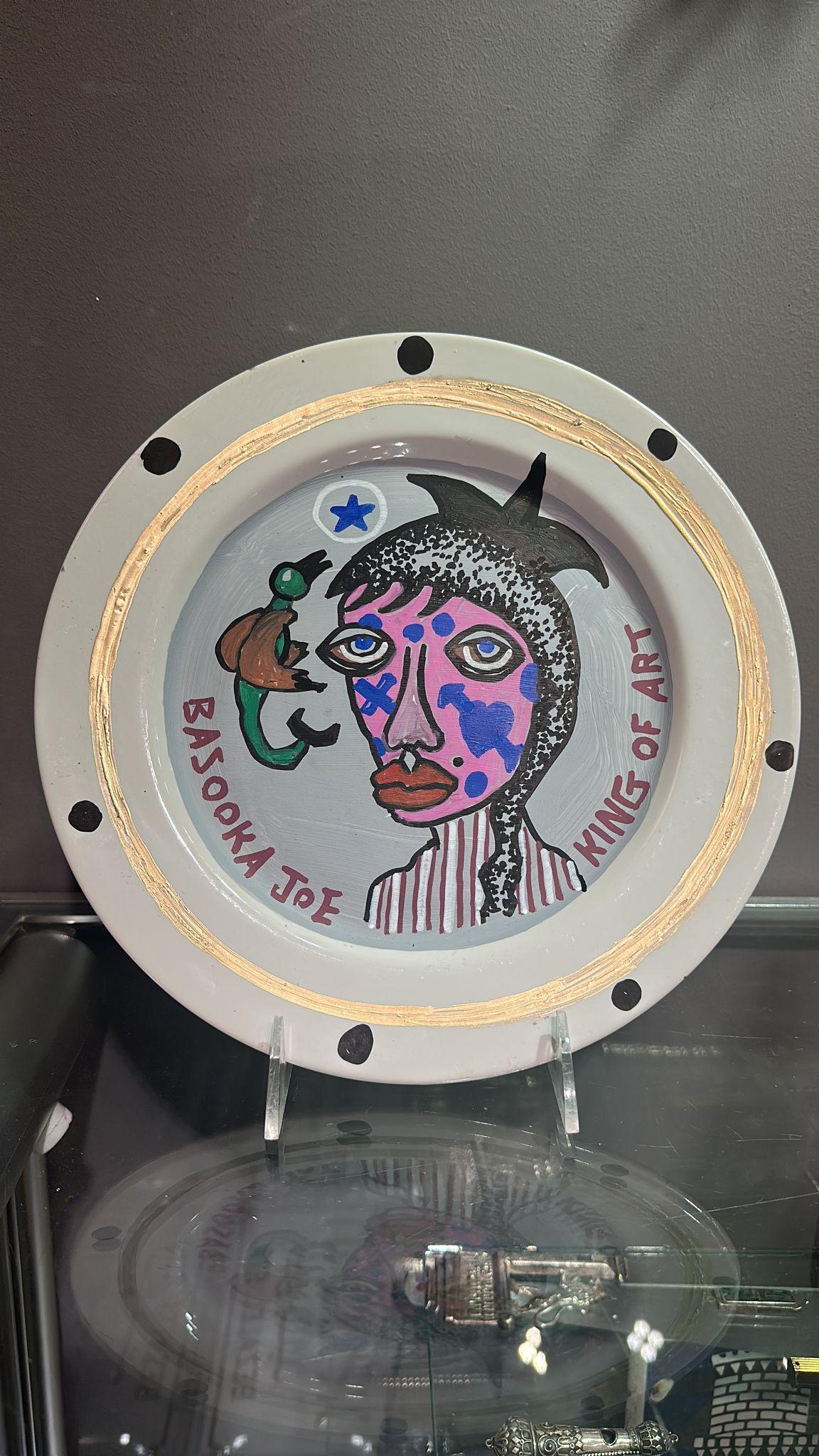 Questo piatto in porcellana dipinto a mano dall'artista israeliano Bazooka Joe presenta un ritratto impressionante ed espressivo realizzato in acrilico. Il soggetto, raffigurato con contorni decisi e colori vivaci, ha un viso rosa ornato da segni